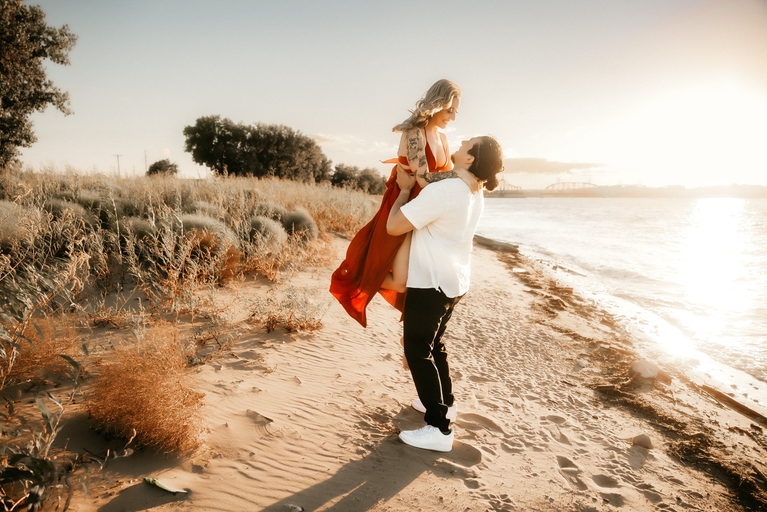 A man lifting a woman in a red dress on a beach during sunset.