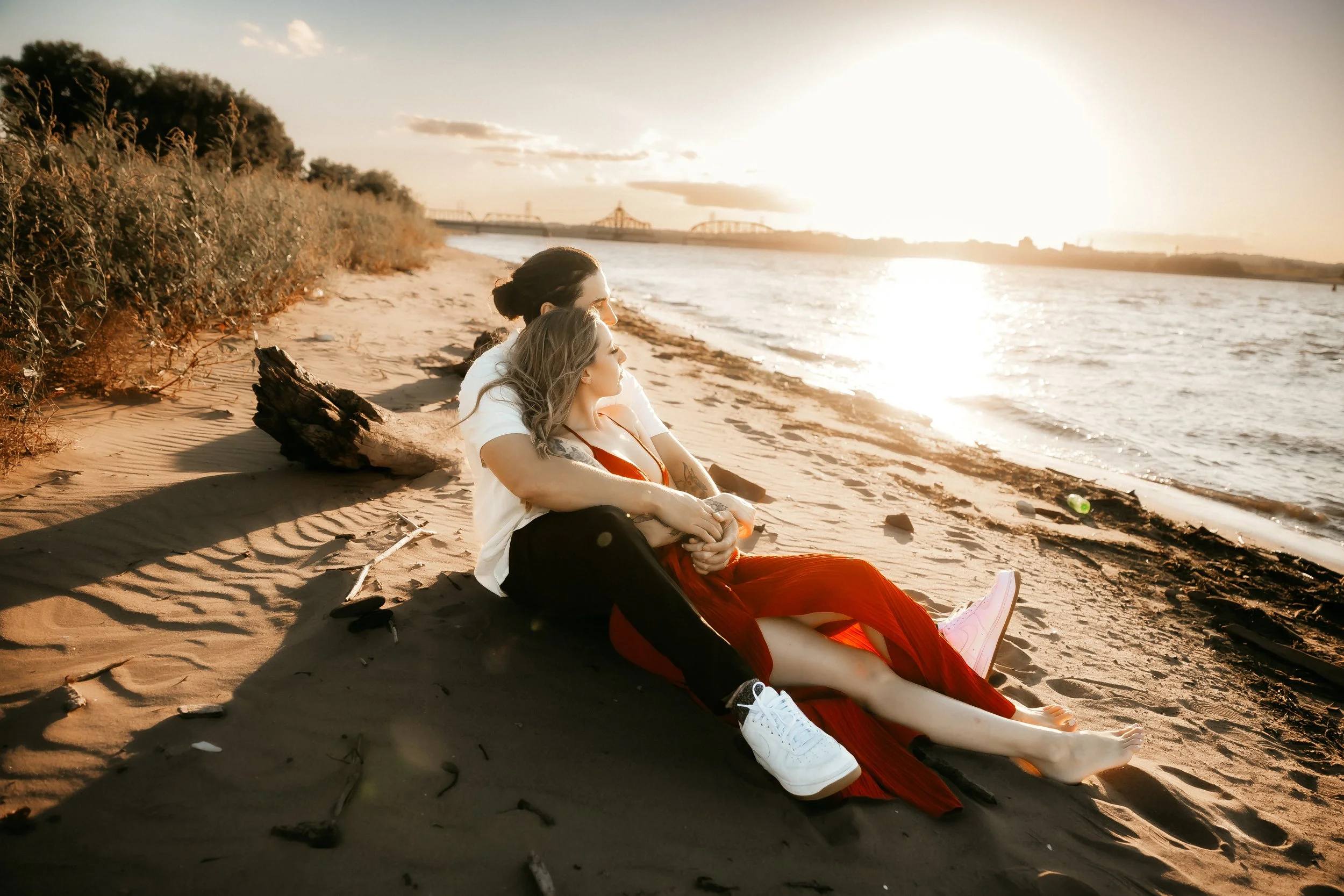 A couple sitting on a sandy beach during sunset, facing the water, with a bridge and sky in the background.