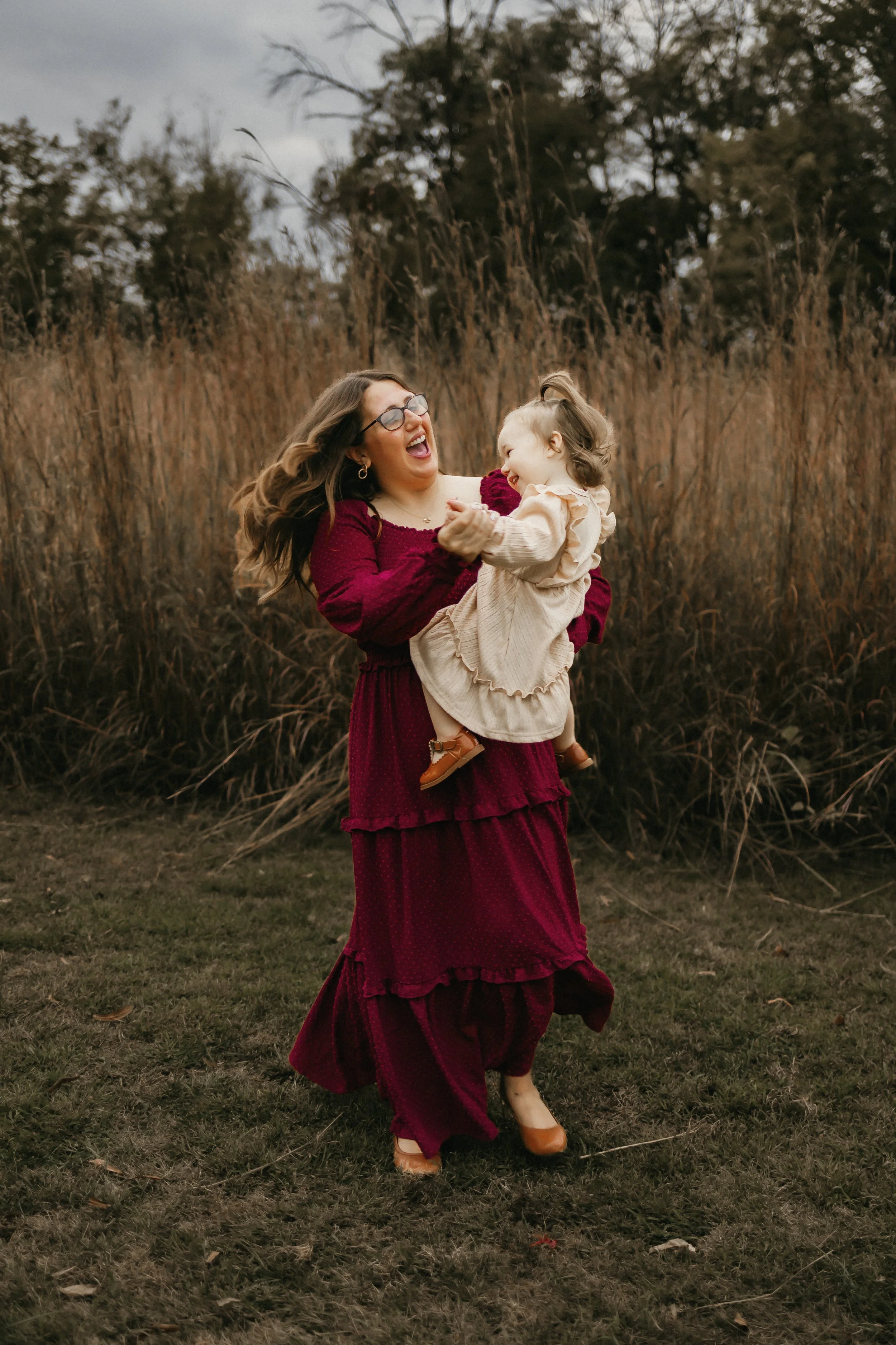 A woman wearing glasses and a long maroon dress is holding a young girl in a beige dress and brown shoes, both smiling and laughing outdoors amidst tall dry grass and trees.