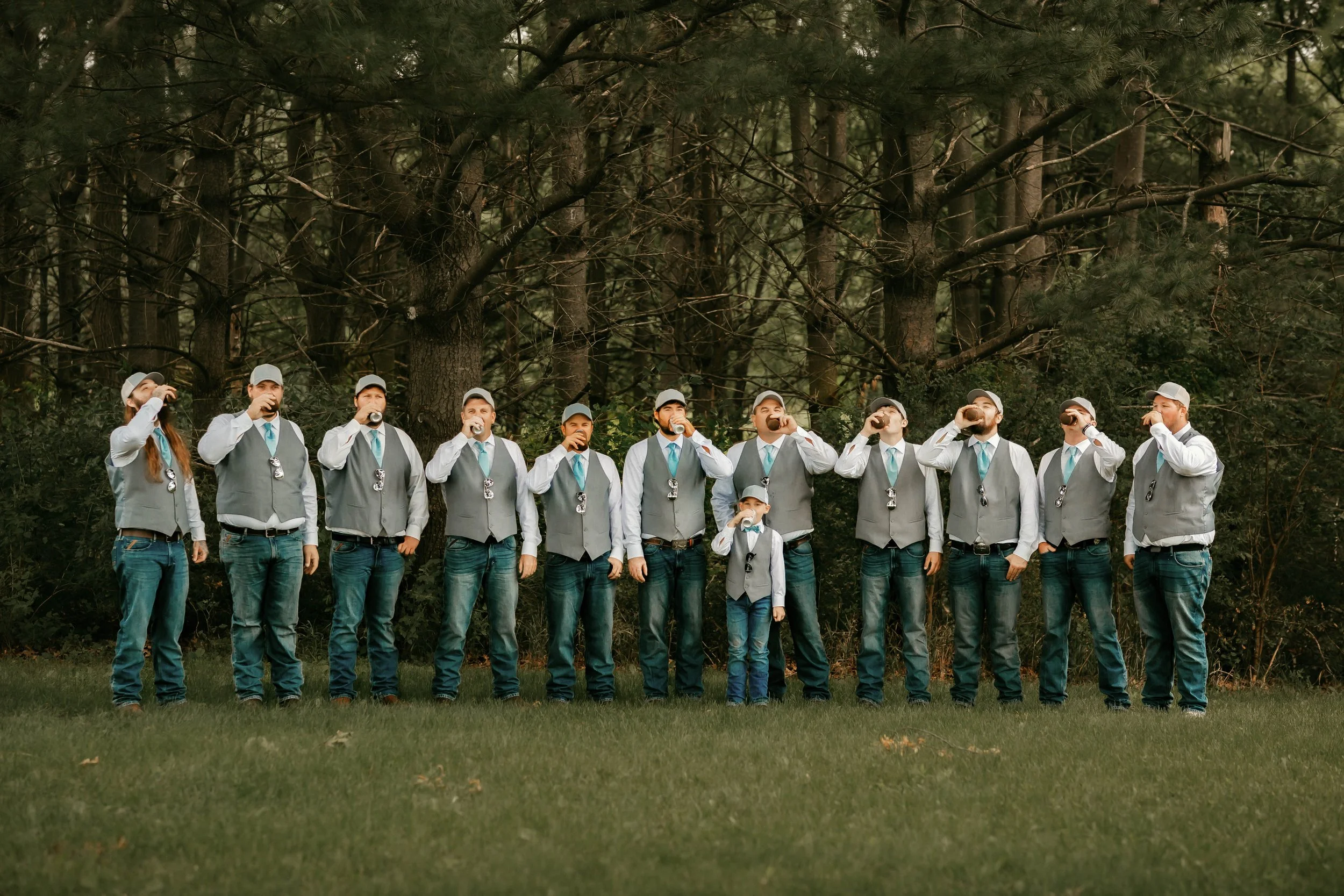 Group of twelve men and one boy dressed in gray vests, white shirts, jeans, and gray caps drinking from bottles outdoors in a forested area.