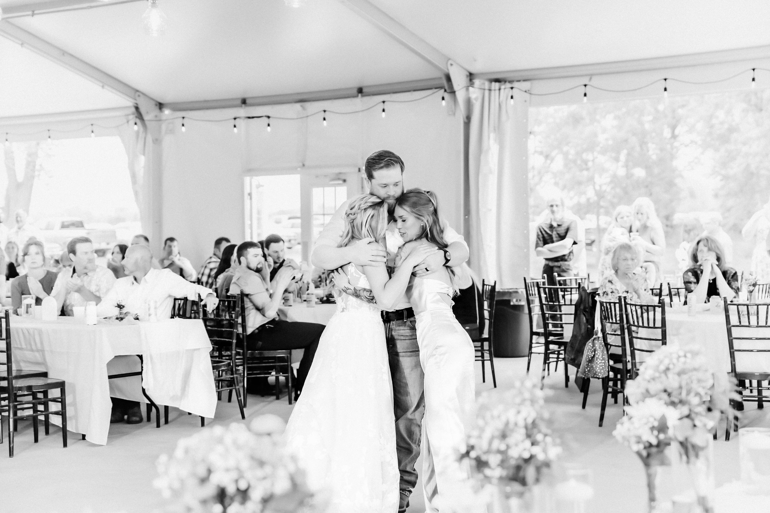 A black-and-white photo of a wedding reception with three people embracing in the center. Guests are sitting at tables in the background, some clapping and others watching. The setting is inside a large tent with string lights.