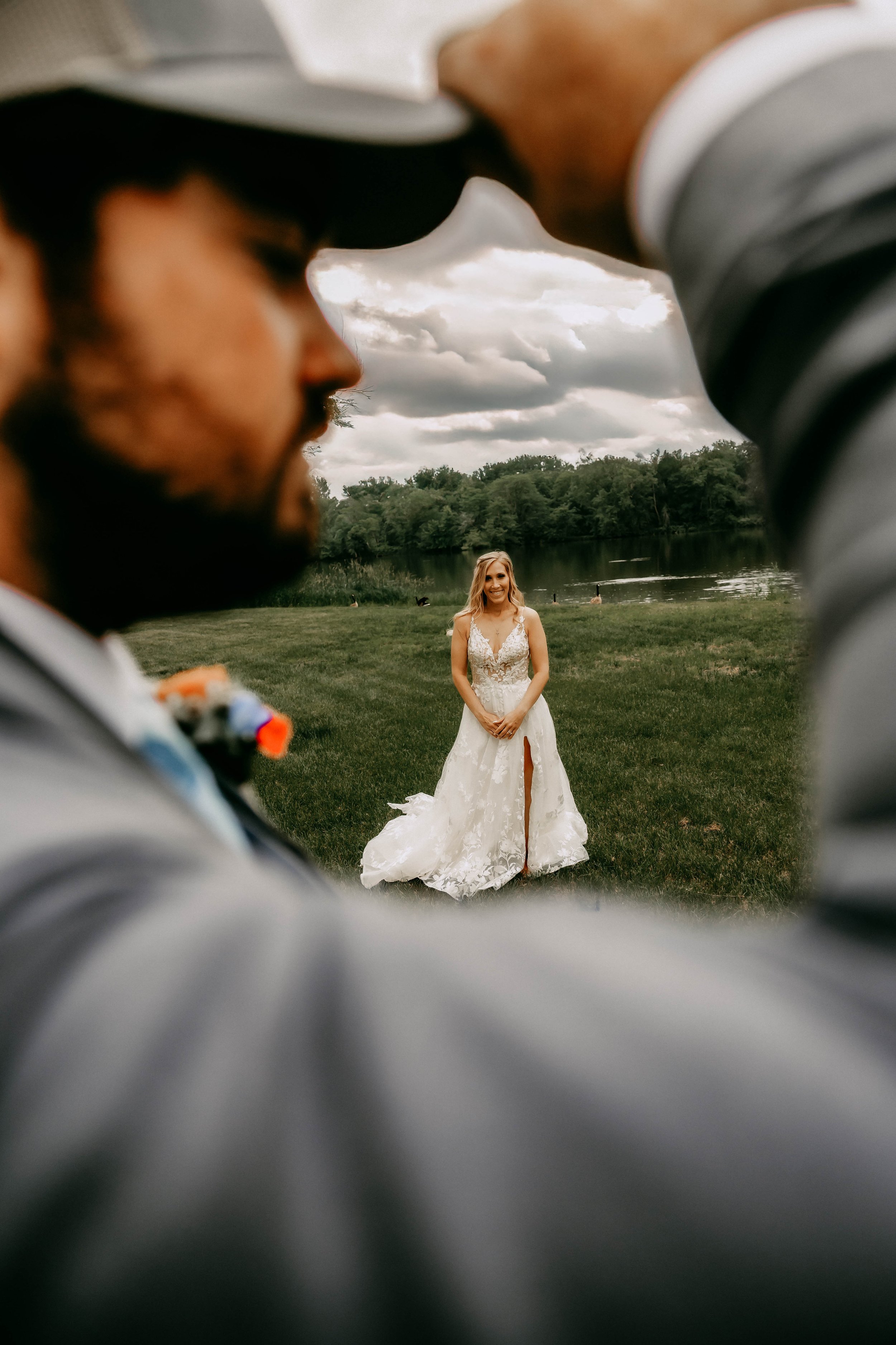 A bride in a white wedding dress standing on grass near a lake, seen through a heart-shaped view formed by two people in suits taking a photo.
