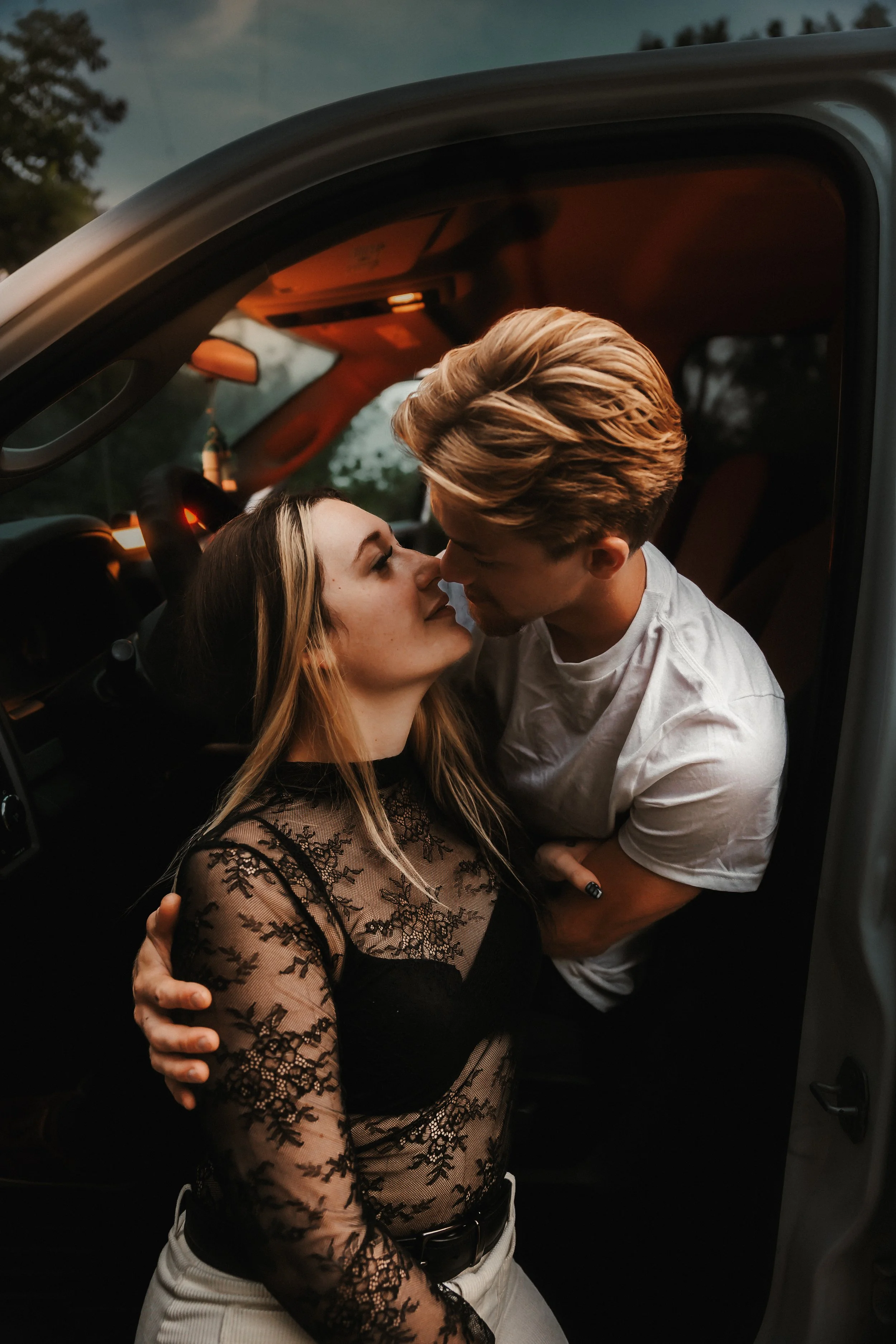 A couple sharing an intimate moment inside a vehicle, close faces, arm around each other, woman in black lace top, man in white t-shirt, evening setting.