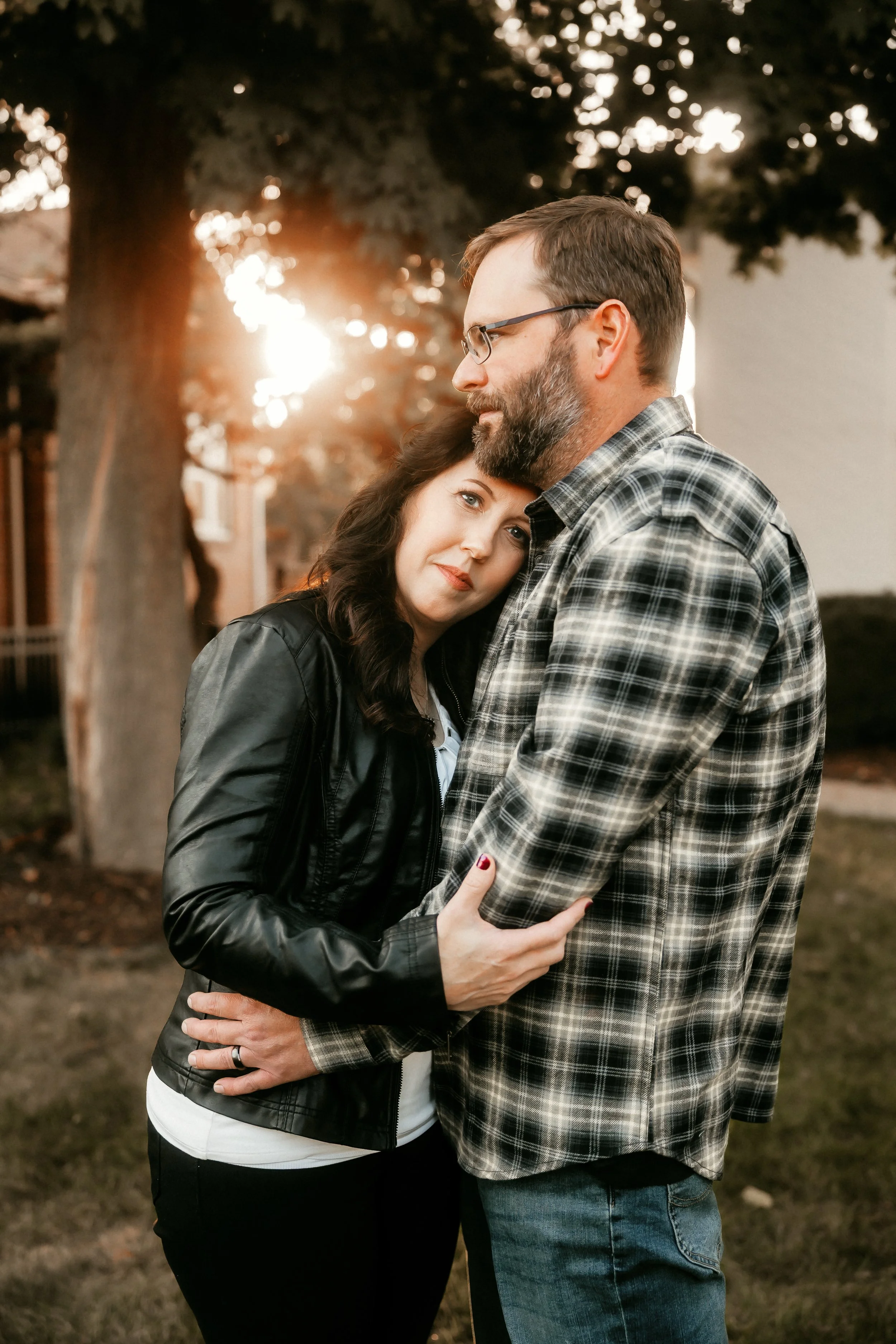 A woman and a man embrace outdoors at sunset, with the woman resting her head on the man's chest and both looking content.