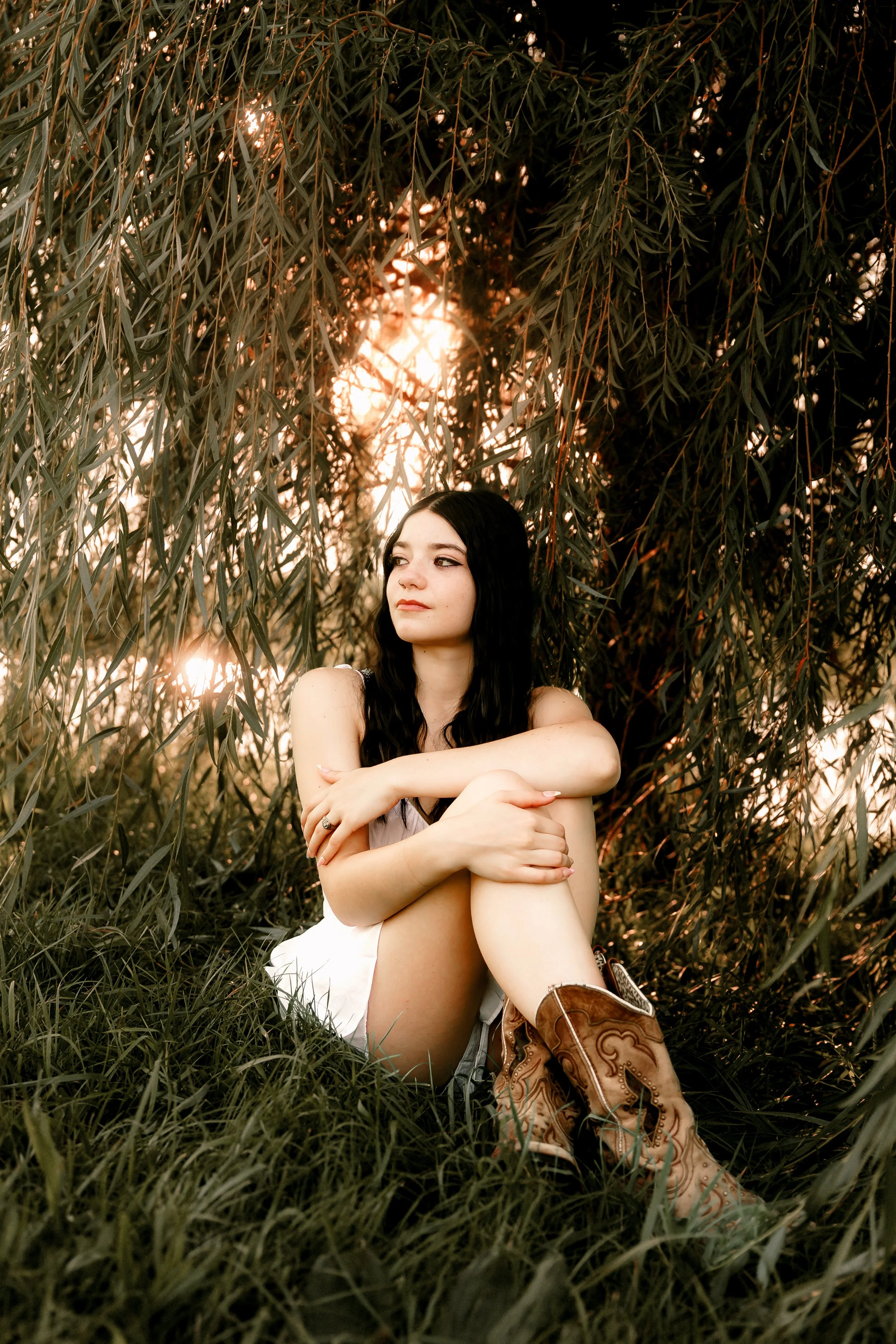 A young woman with black hair and light skin sits in grass under a tree, wearing a white dress and cowboy boots, with sunlight filtering through the leaves.