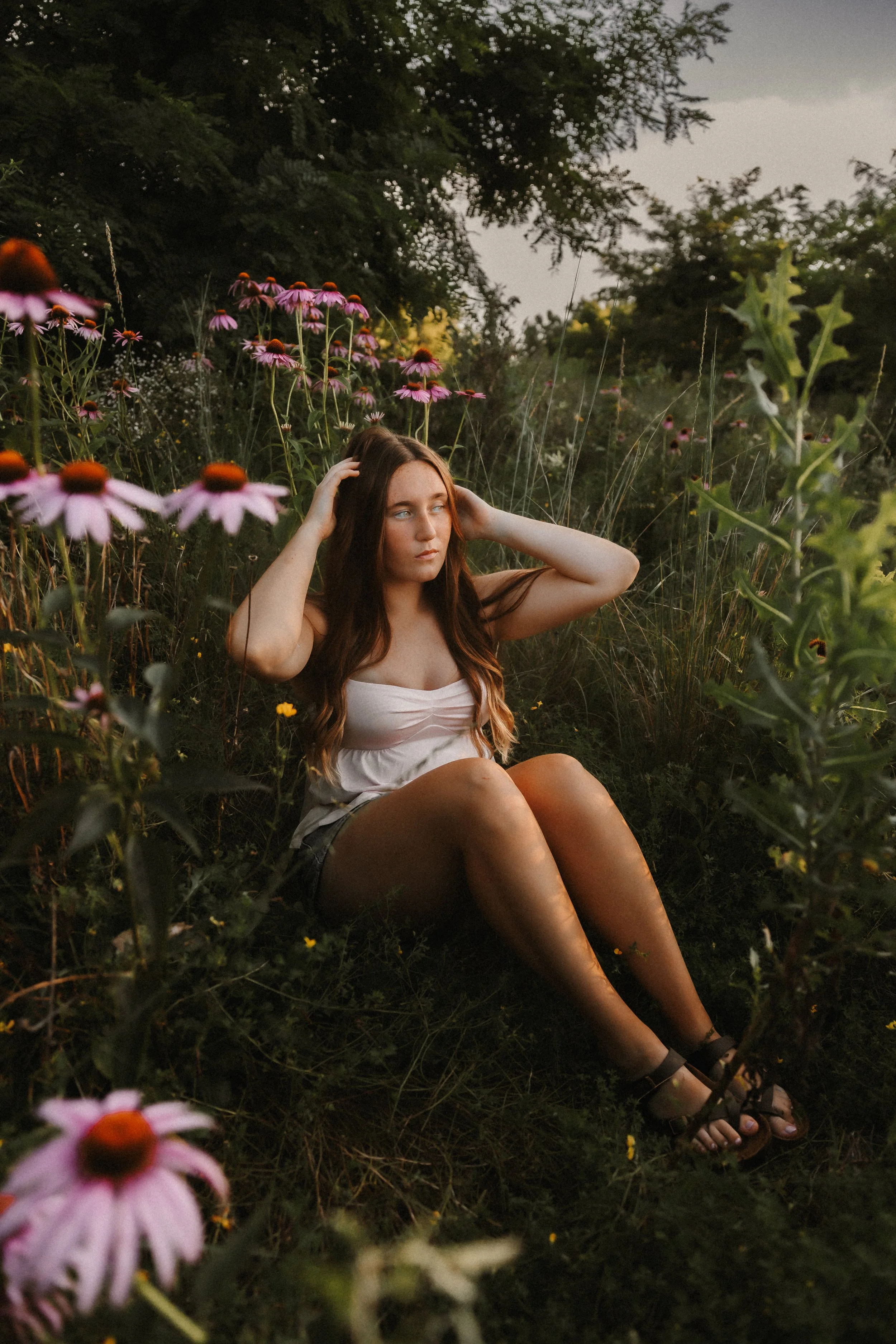 A young woman with long wavy brown hair sitting in a field of wildflowers, wearing a white top and dark shorts, with her hands touching her head, surrounded by pink coneflowers and tall grass.