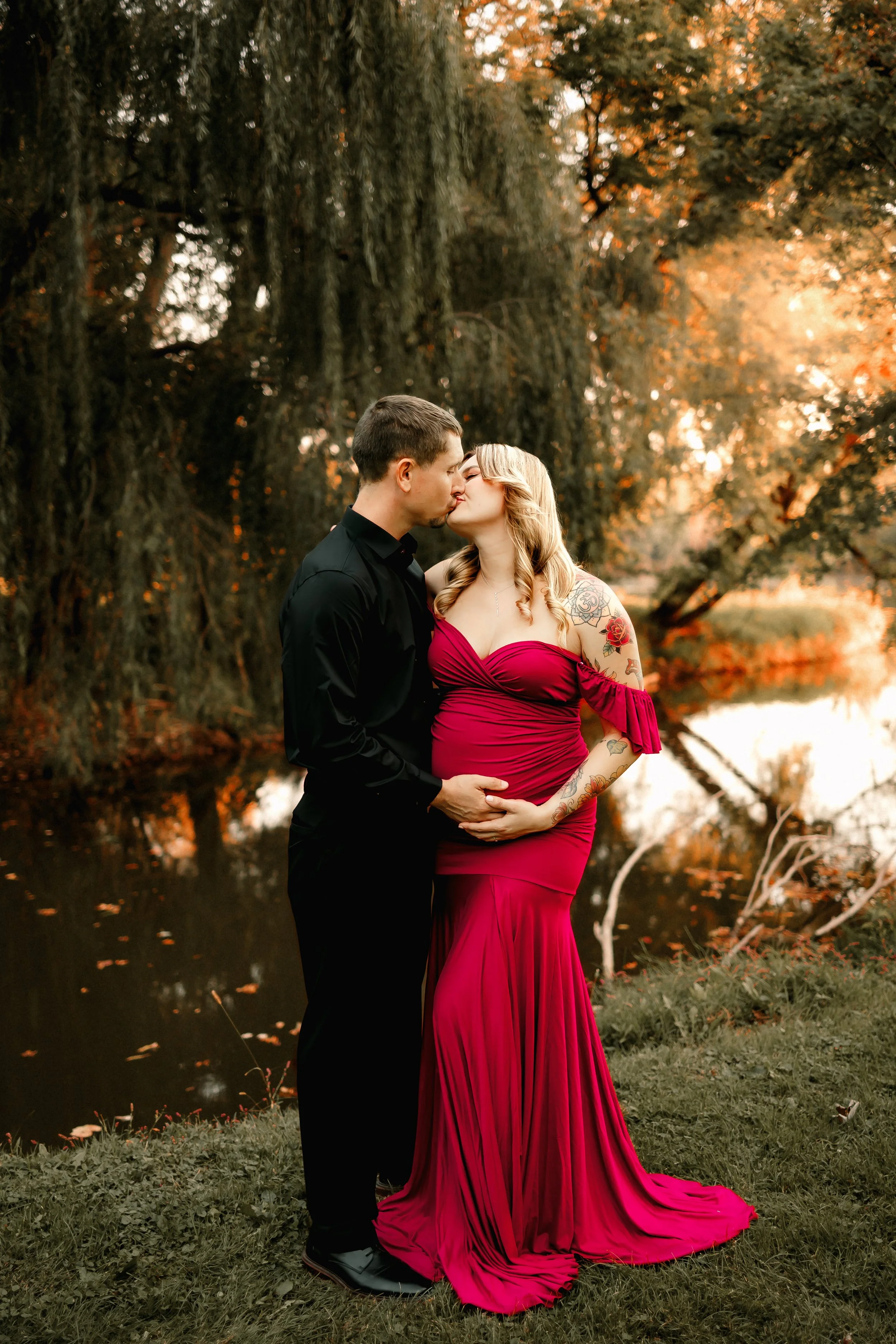 A couple kissing outdoors by a pond during sunset, the woman is wearing a red gown and has tattoos on her arm, and the man is in a black shirt and pants.