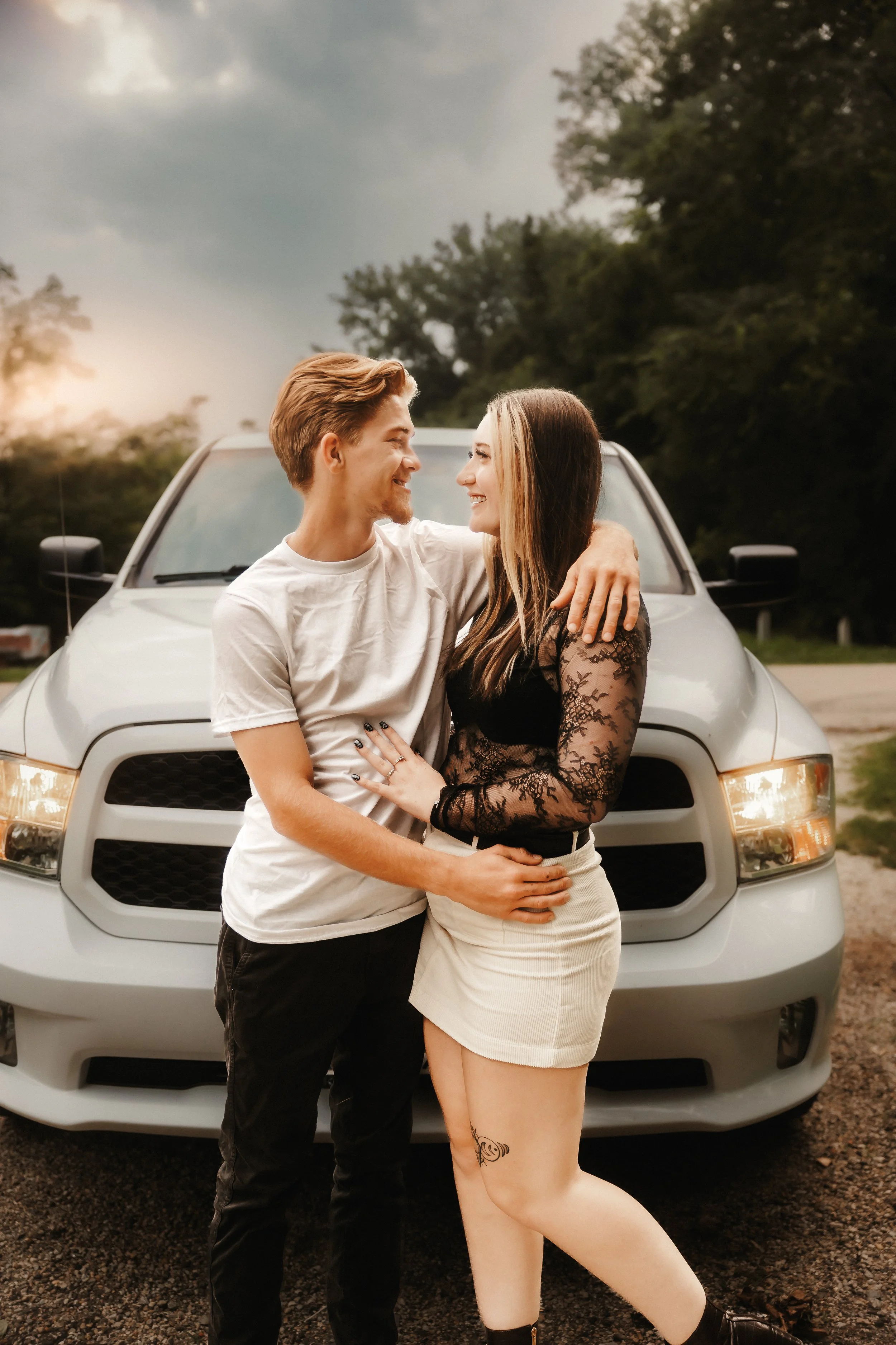 A young couple smiling and embracing in front of a silver SUV on a rural road during sunset.