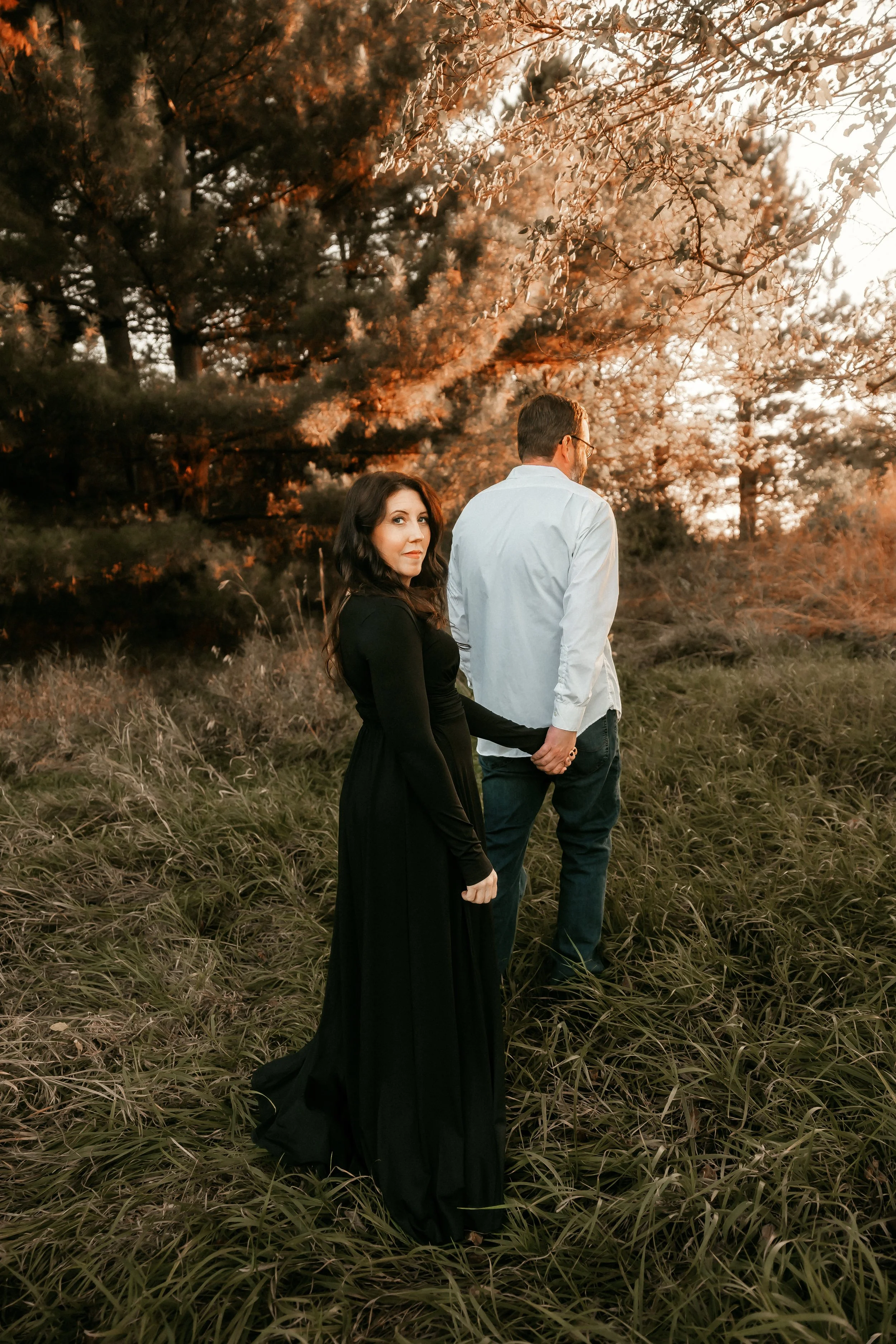 A woman in a long black dress holding hands with a man in a white shirt and jeans, standing outdoors in a grassy area with trees and an orange sunset in the background.
