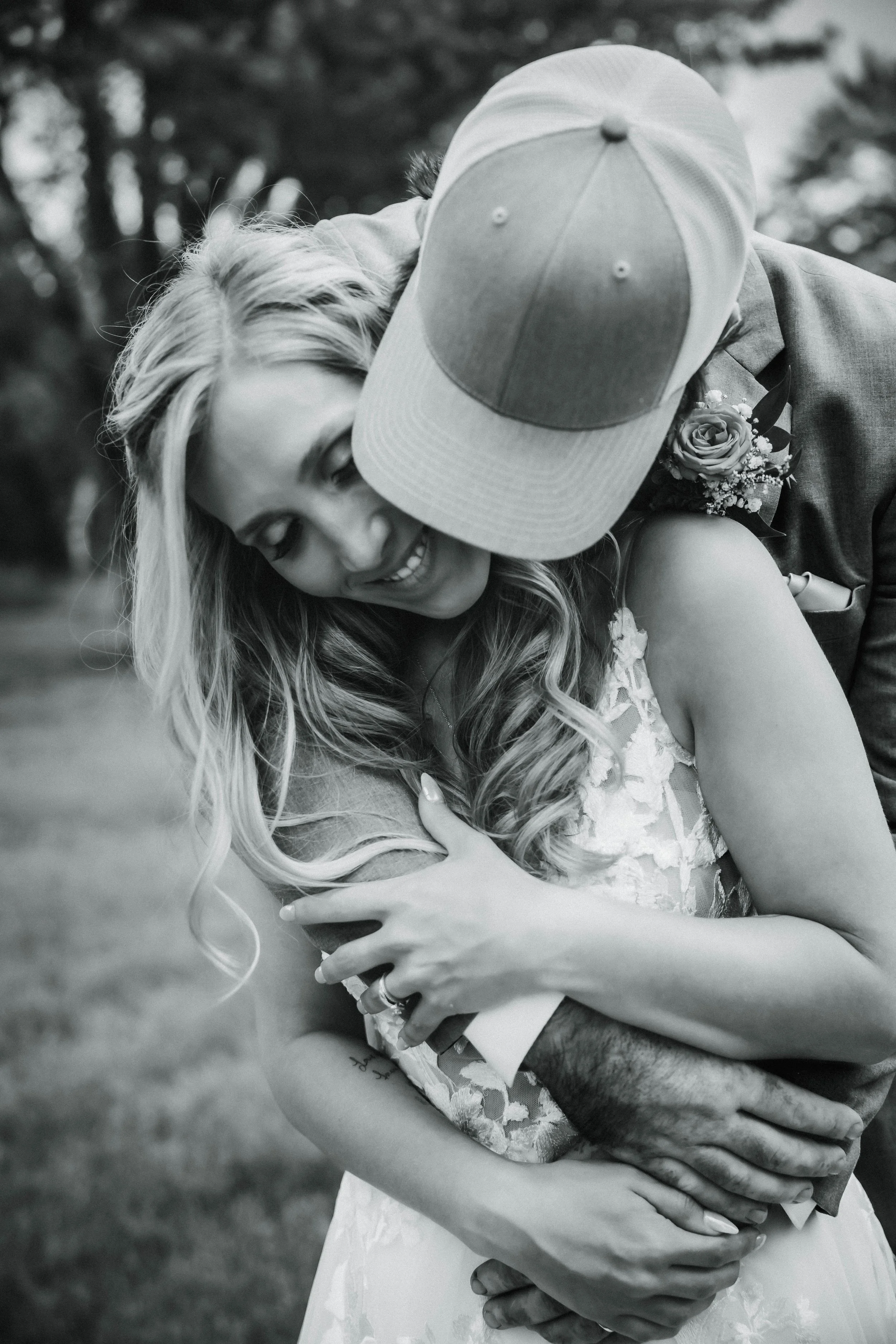 A couple embracing outdoors, the woman smiling with eyes closed, wearing a white dress, the man with a baseball cap and a boutonniere, holding her from behind.