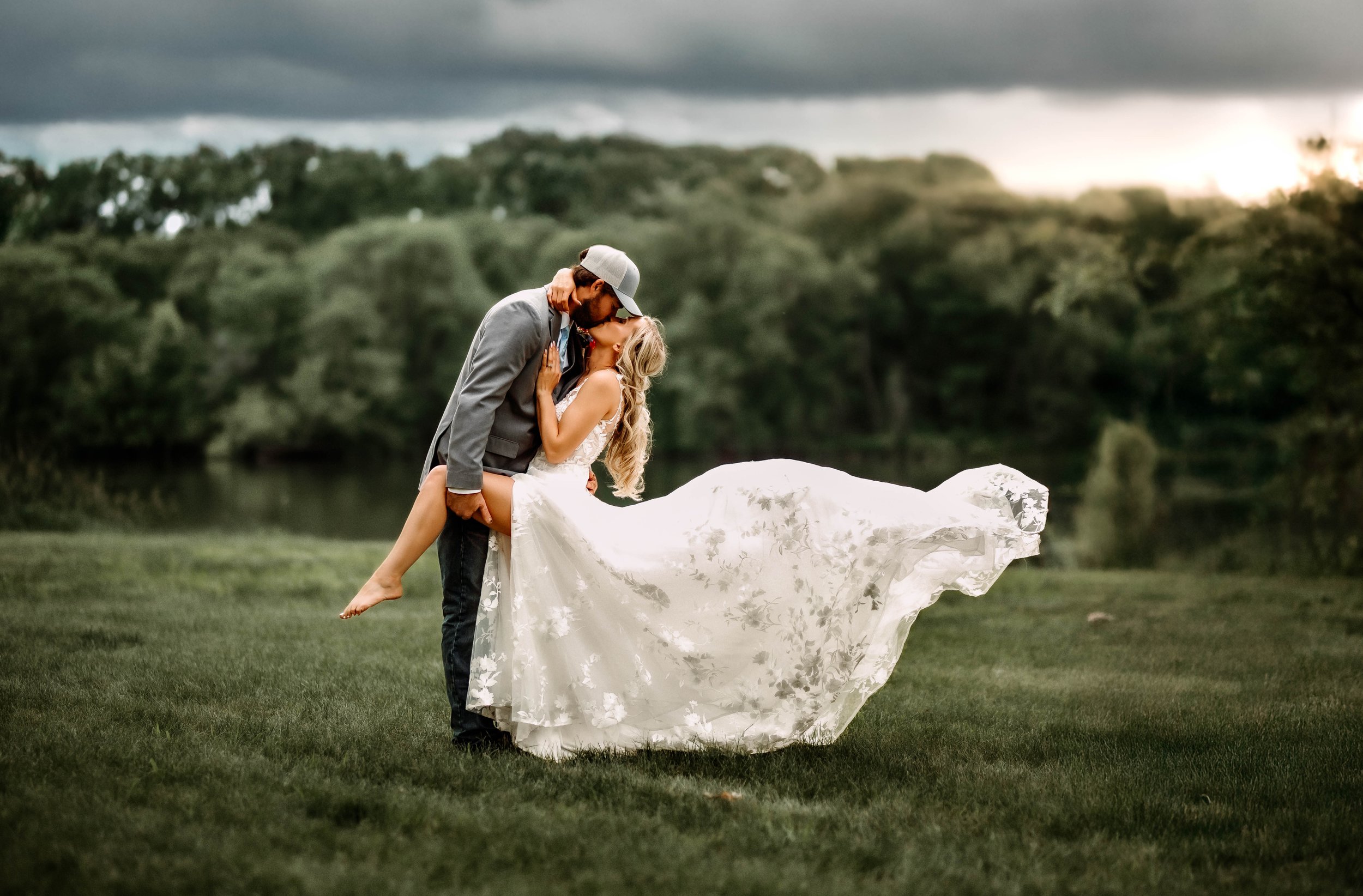 A couple kissing outdoors, the man lifting the woman in a white wedding dress with a lakeside background and cloudy sky.