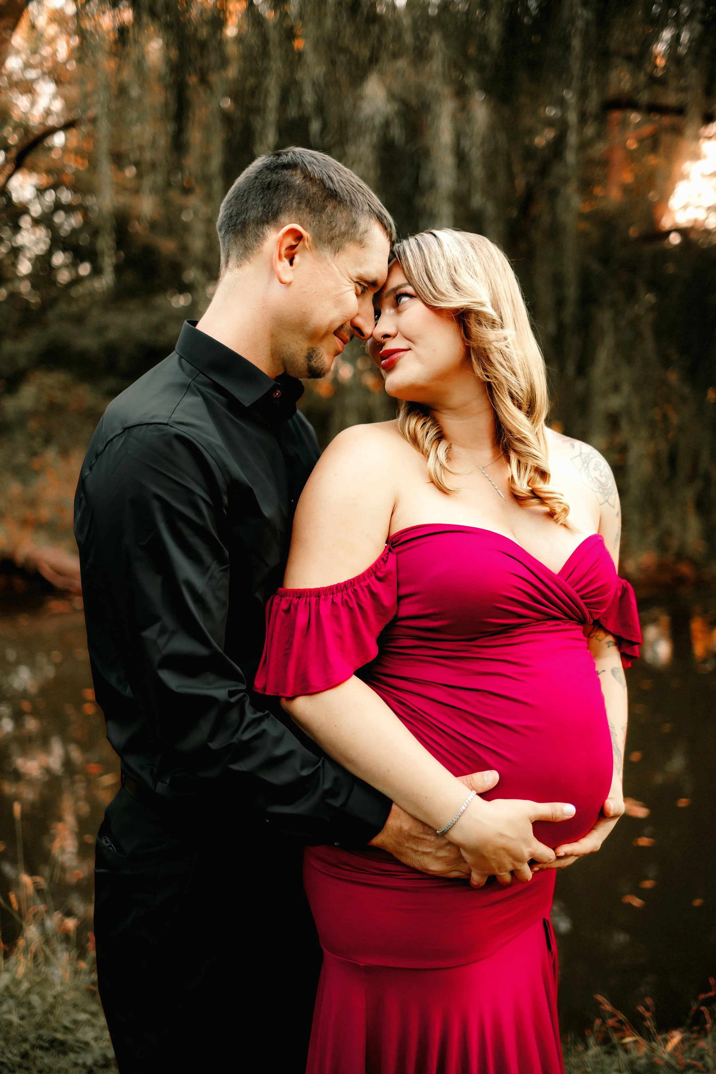 A pregnant woman in a red off-the-shoulder dress with wavy blonde hair, standing outdoors by a body of water, with a man in a black shirt holding her belly and touching her hand, both leaning in with heads touching, in a romantic pose in a natural setting with trees in the background.