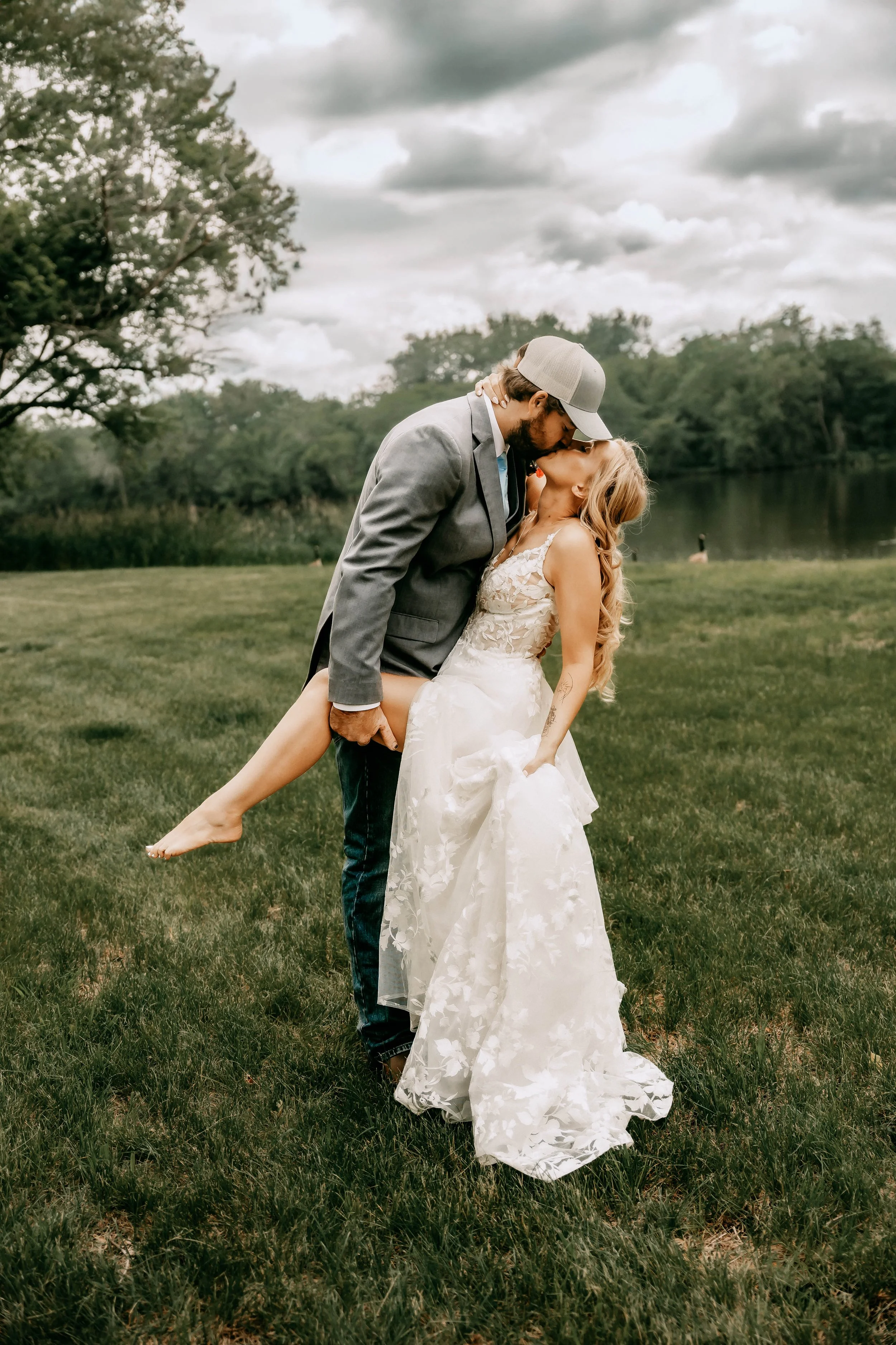 Couple in wedding attire sharing a kiss outdoors near a lake, with a man in a gray suit and cap holding a woman in a white lace wedding dress, as they embrace on a grassy area.