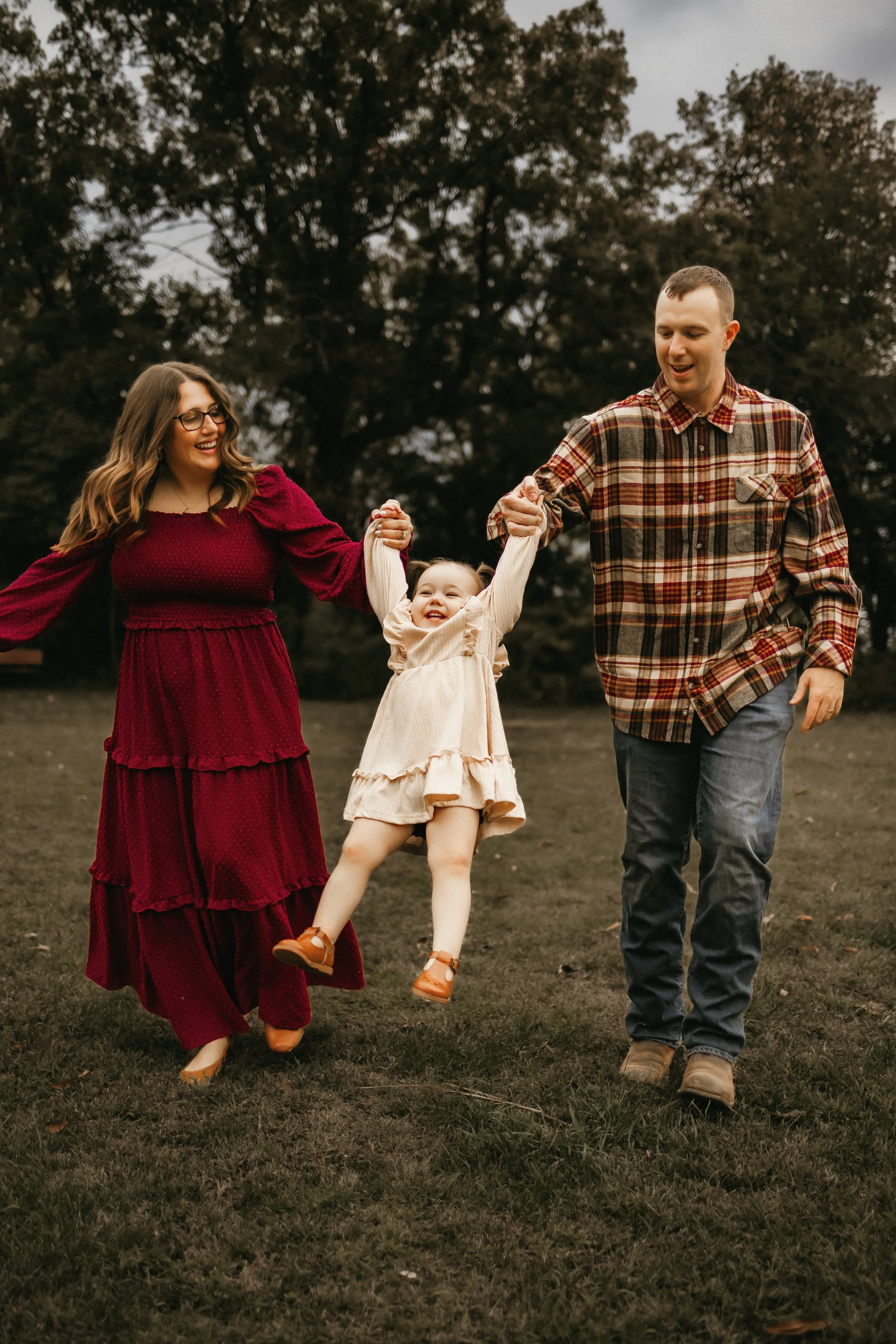 A family of three enjoying a playful moment outdoors on a cloudy day, with a woman wearing a red dress, a man in a plaid shirt, and a young girl in a beige dress swinging between them.