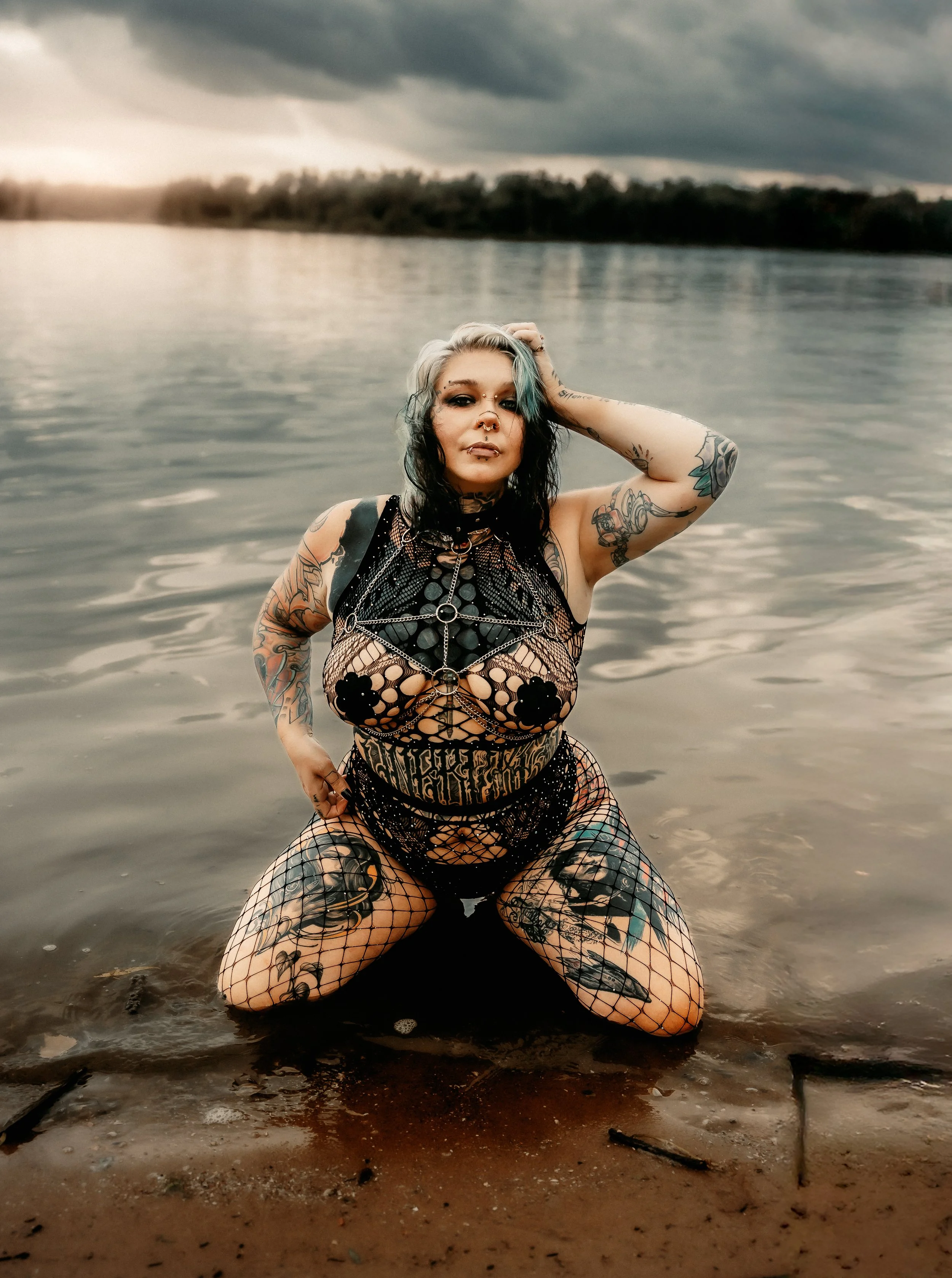 A tattooed woman kneeling in shallow water at the edge of a lake or river, with cloudy skies and trees in the background. At Buffalo Beach in Buffalo Iowa