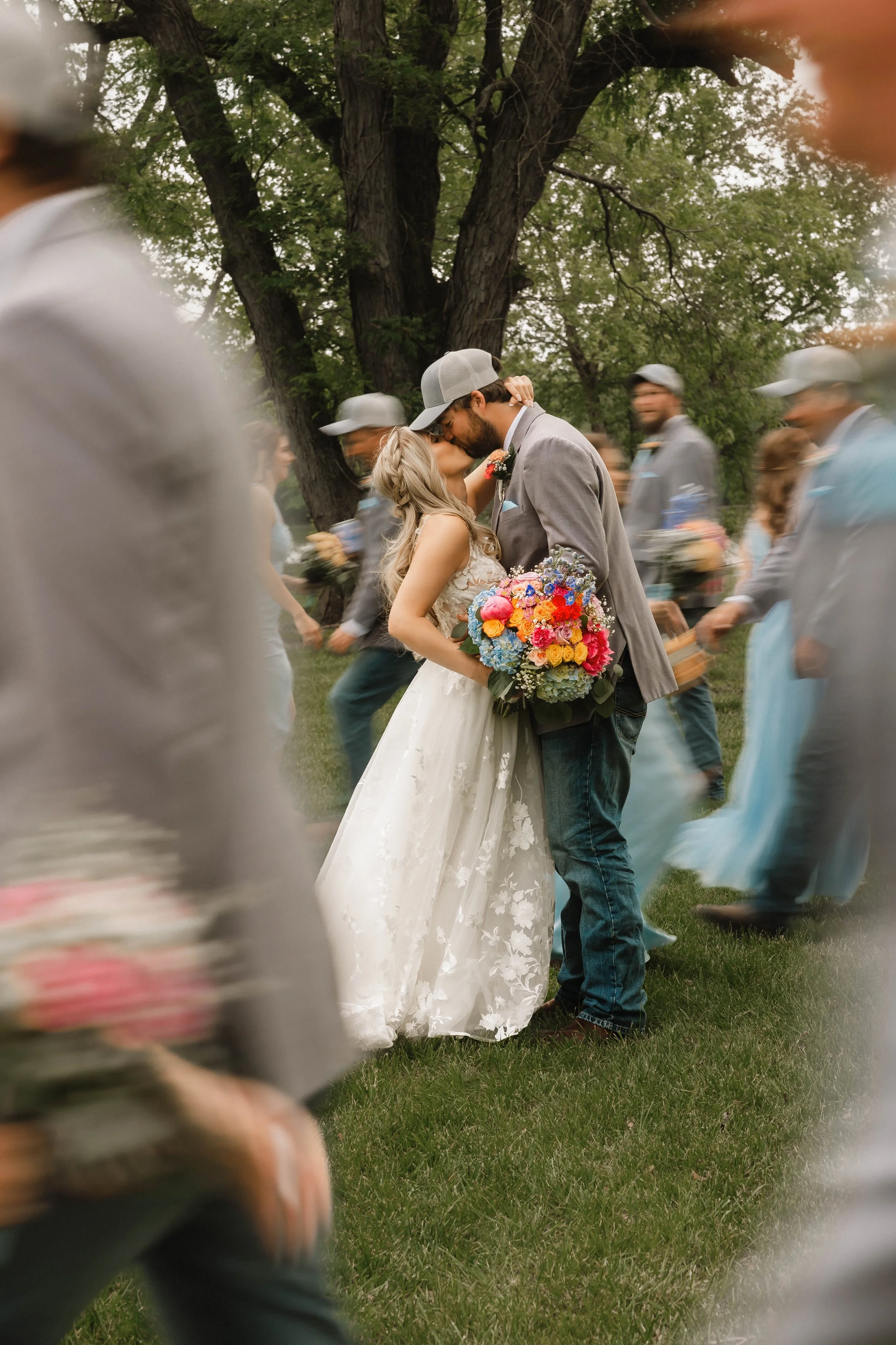 A wedding couple kissing outdoors surrounded by friends and family, with the bride holding a colorful bouquet of flowers, and groom wearing a gray suit jacket and cap, under a large tree.