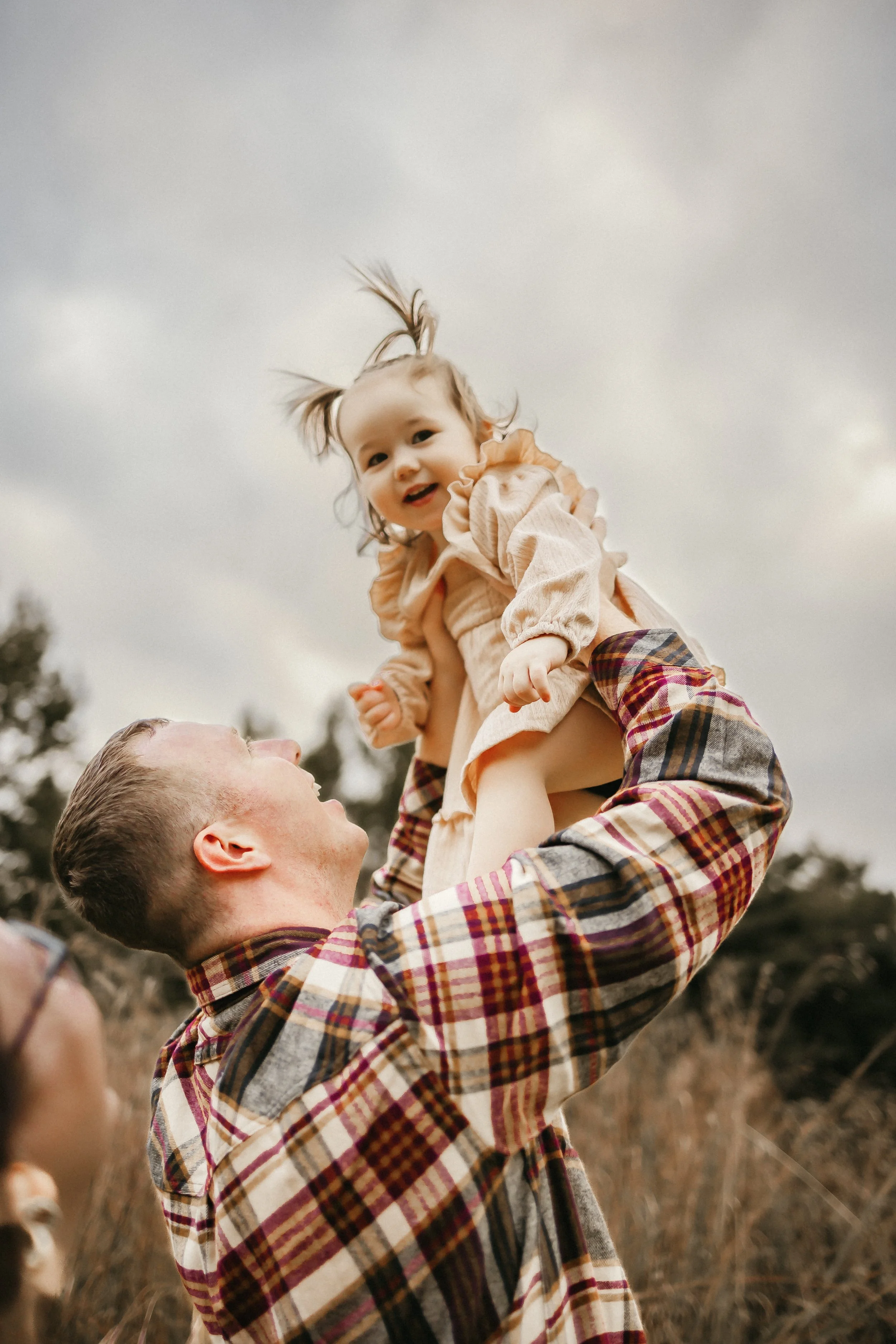 A man lifting a young girl into the air outdoors on a cloudy day, with the girl smiling and playing, surrounded by tall grass and trees.