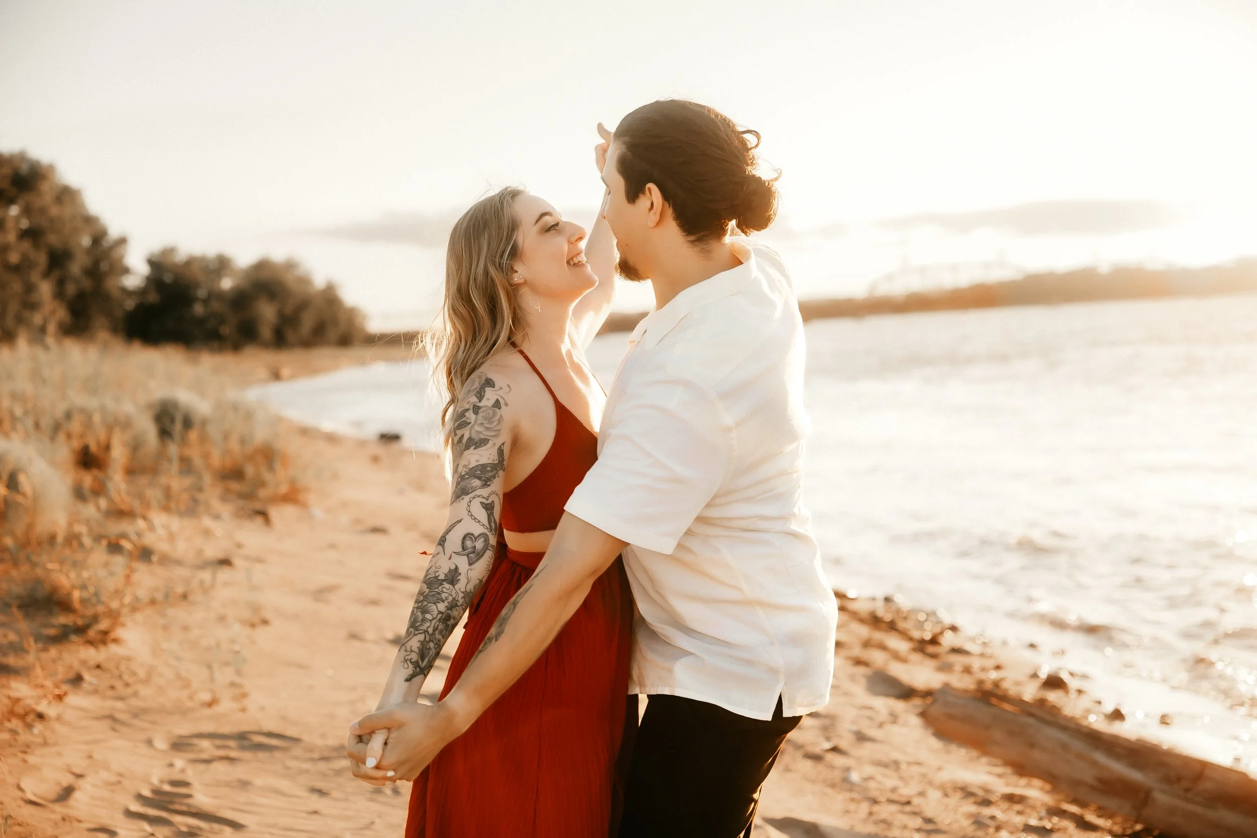 A couple standing on a sandy beach holding hands and looking at each other at sunset.