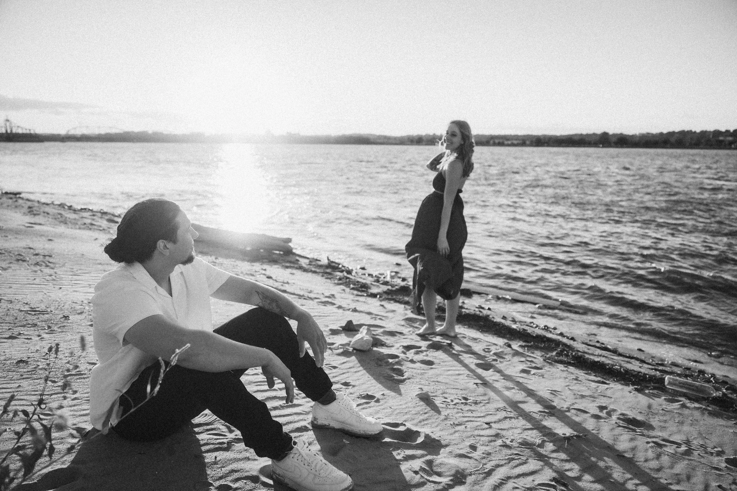 A black-and-white photo of two people by a river, with a woman in a dress standing in the water and a man sitting on the sandy shore looking at her, during sunset.