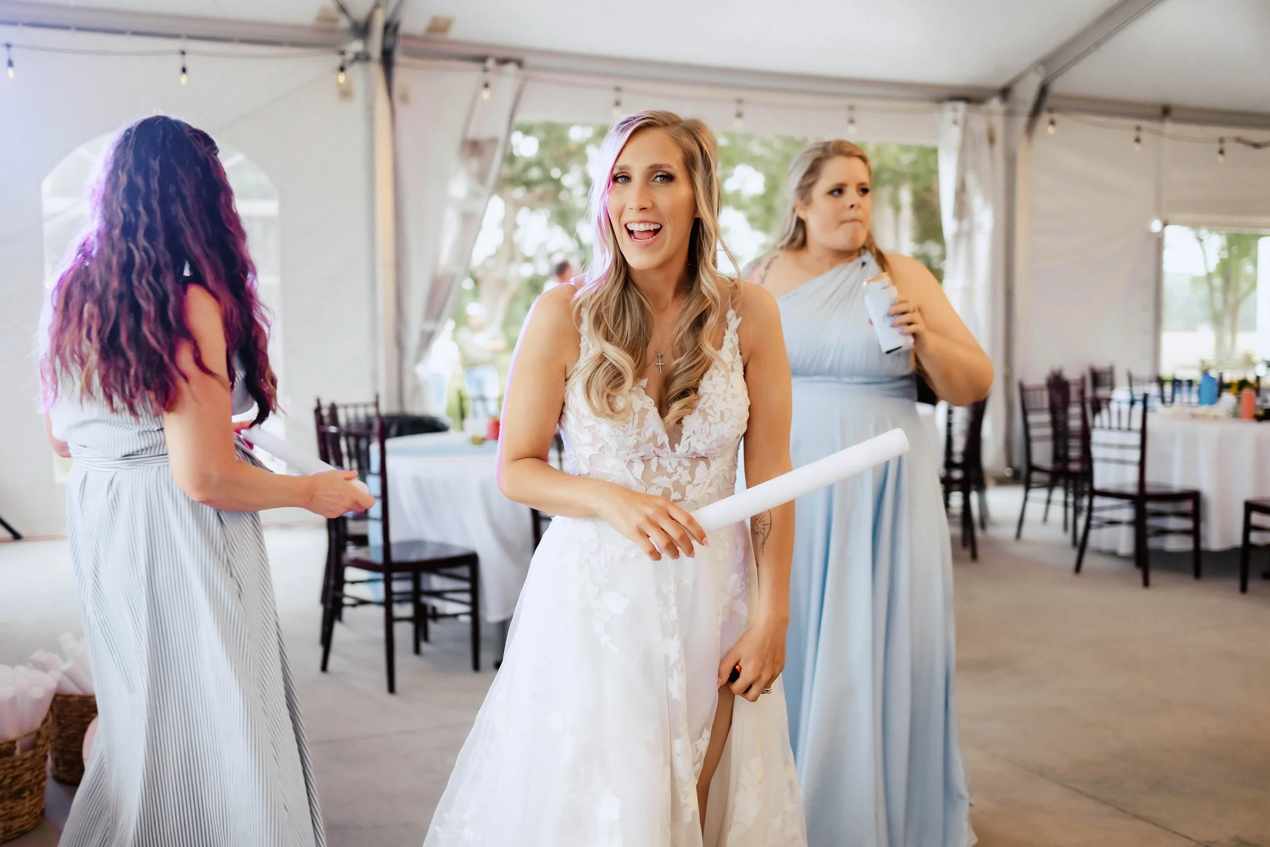 A bride wearing a white lace wedding dress, smiling and holding a rolled-up paper, standing inside a decorated event tent with other women around her.