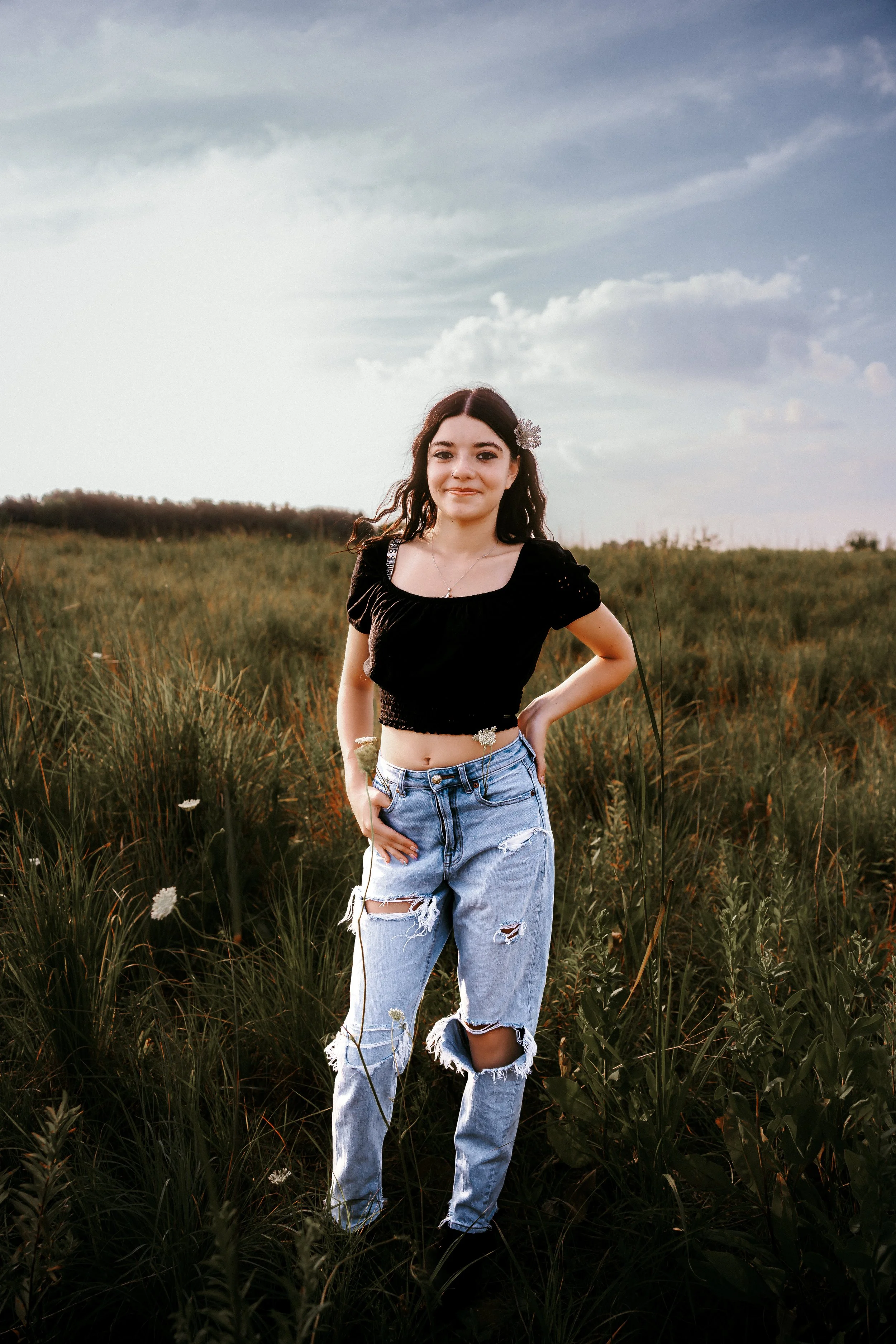 Young woman standing in a grassy field during sunset, wearing a black crop top and ripped jeans, with a flower in her hair.