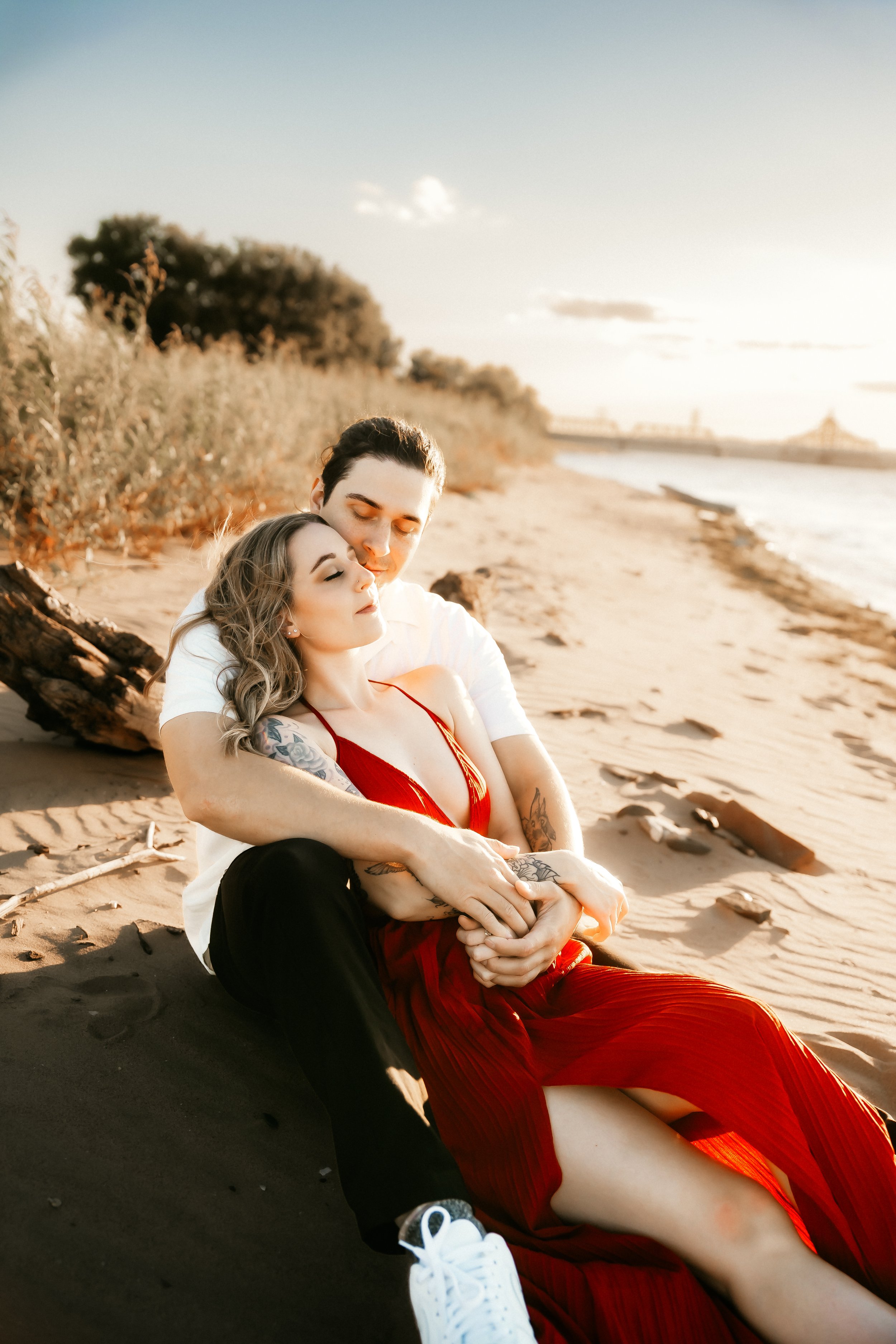 A couple sitting on the beach, primarily in the sunlight, with the woman in a red dress and the man in a white shirt and black pants, embracing each other, surrounded by driftwood and sand, with water and sky in the background.