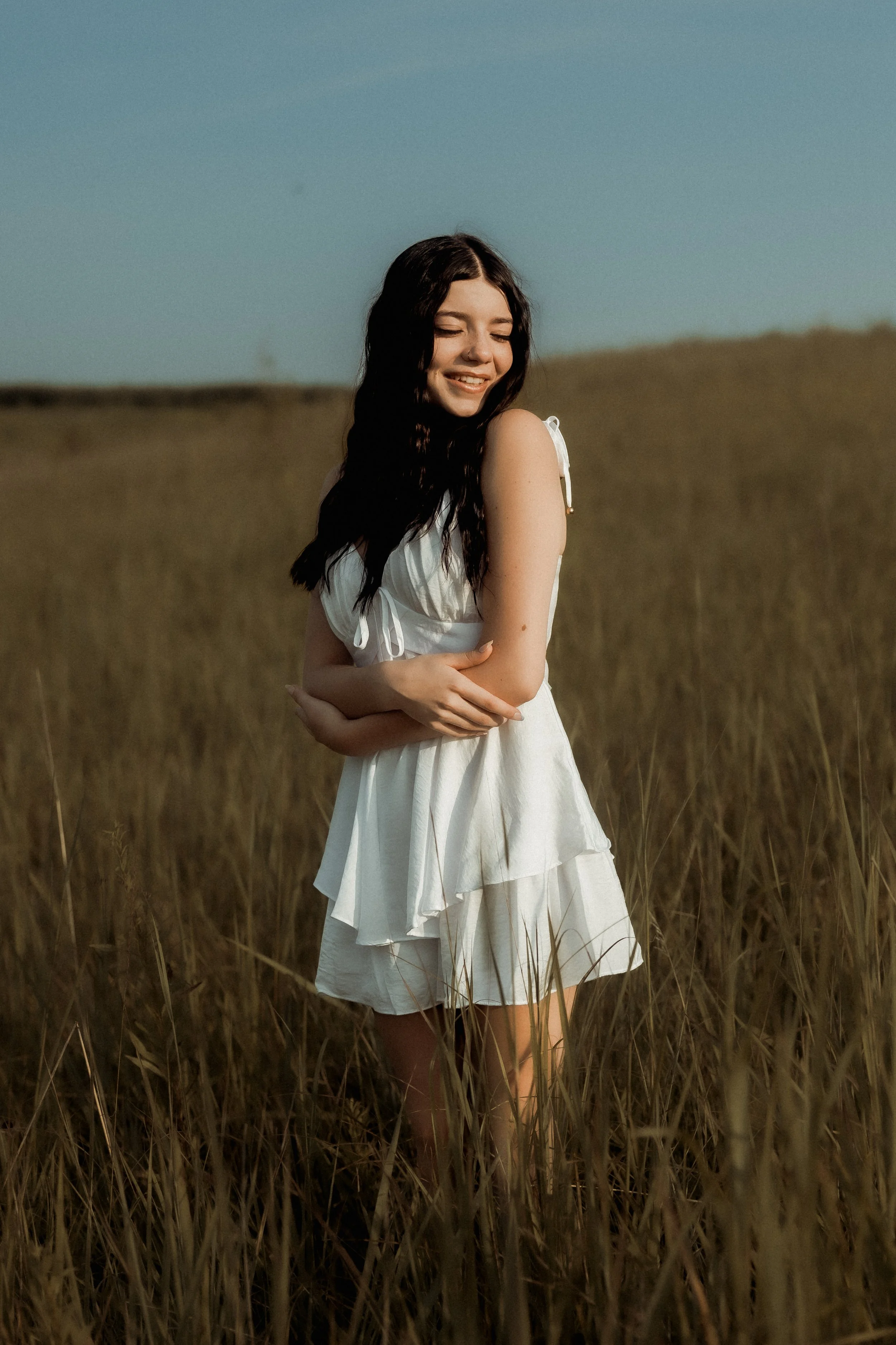 A young woman in a white dress standing in a field of tall grass, smiling with her eyes closed.