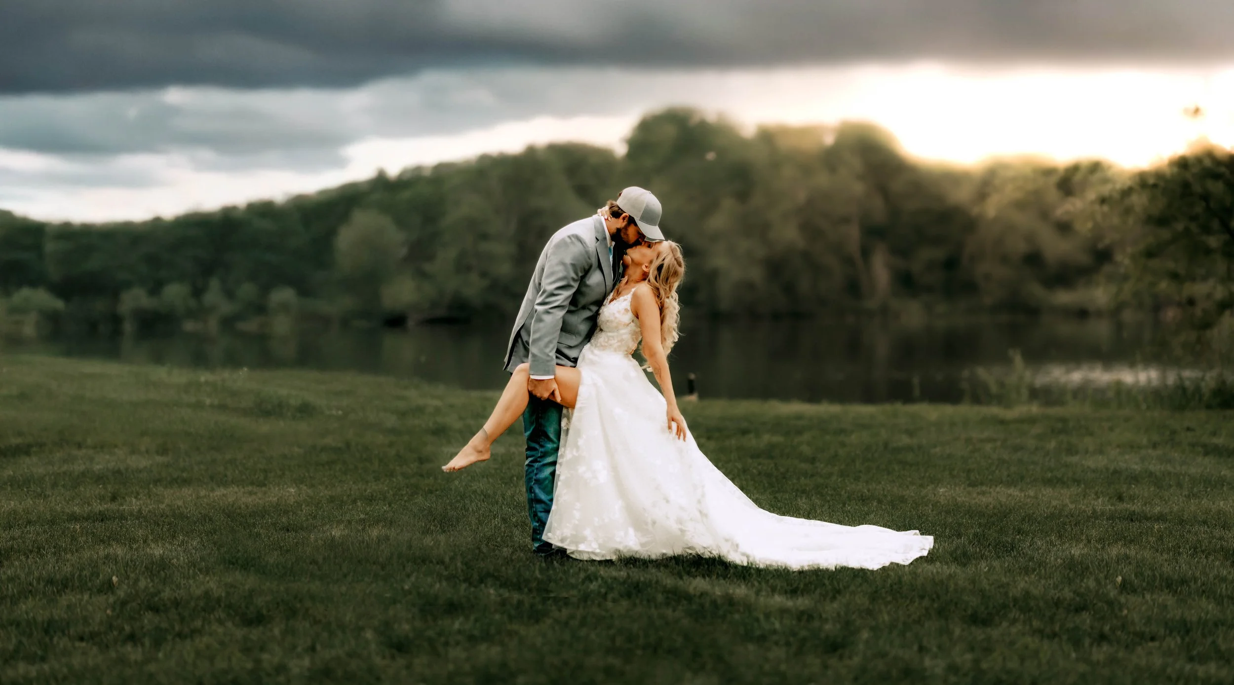 A bride and groom share a kiss outdoors by a lake at sunset, with the groom lifting the bride who is in a white wedding dress.