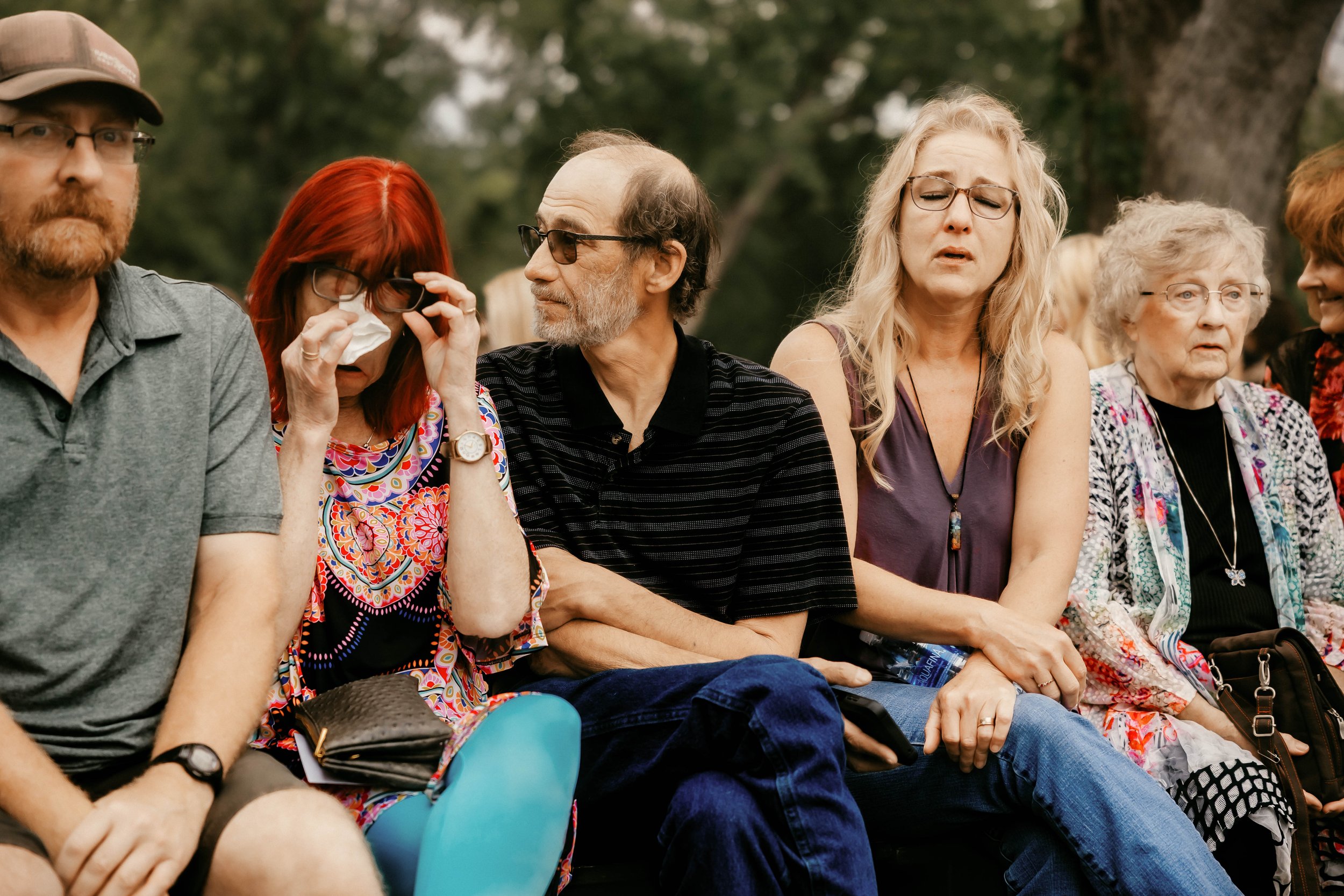 A group of people sitting outdoors, some appear somber or emotional, at what might be a memorial or gathering.