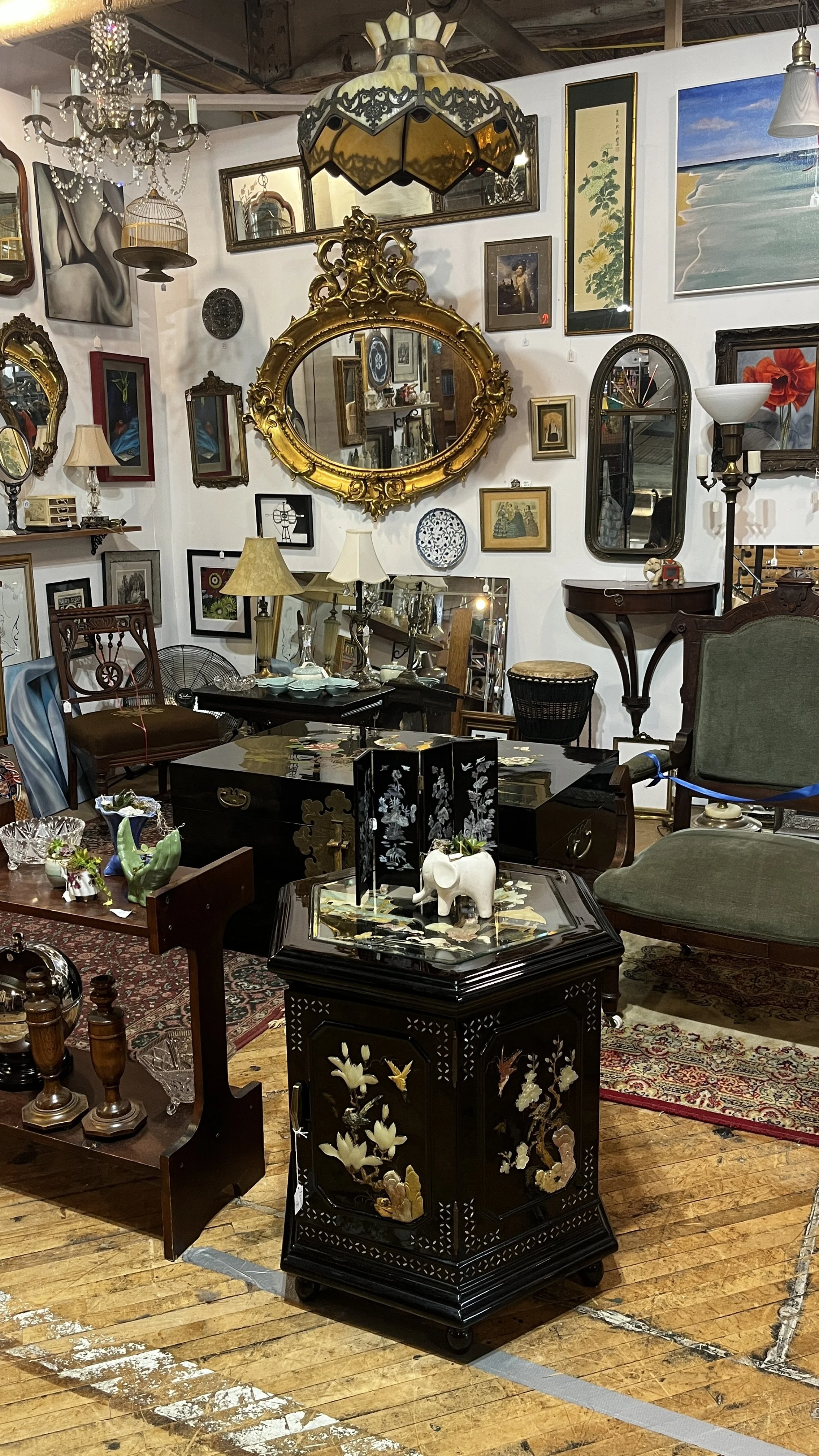 A small antique shop filled with framed artwork on the walls, vintage lamps, mirrors, and decorative items including a black bust, glass vases, and framed pictures. The space has a cozy, cluttered feel with furniture pieces and various collectibles.
