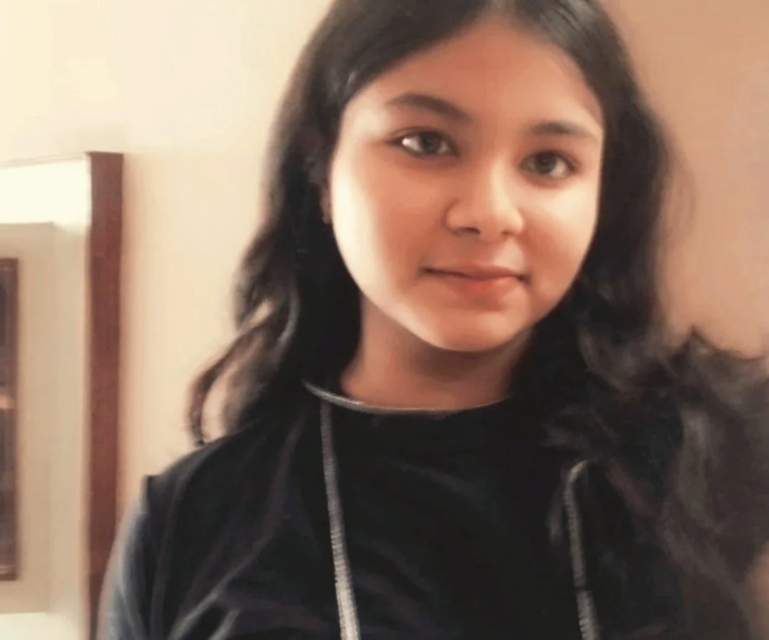 A young woman with dark hair and a slight smile, wearing a black top or jacket, standing indoors in front of a wall with a mirror and a doorframe.