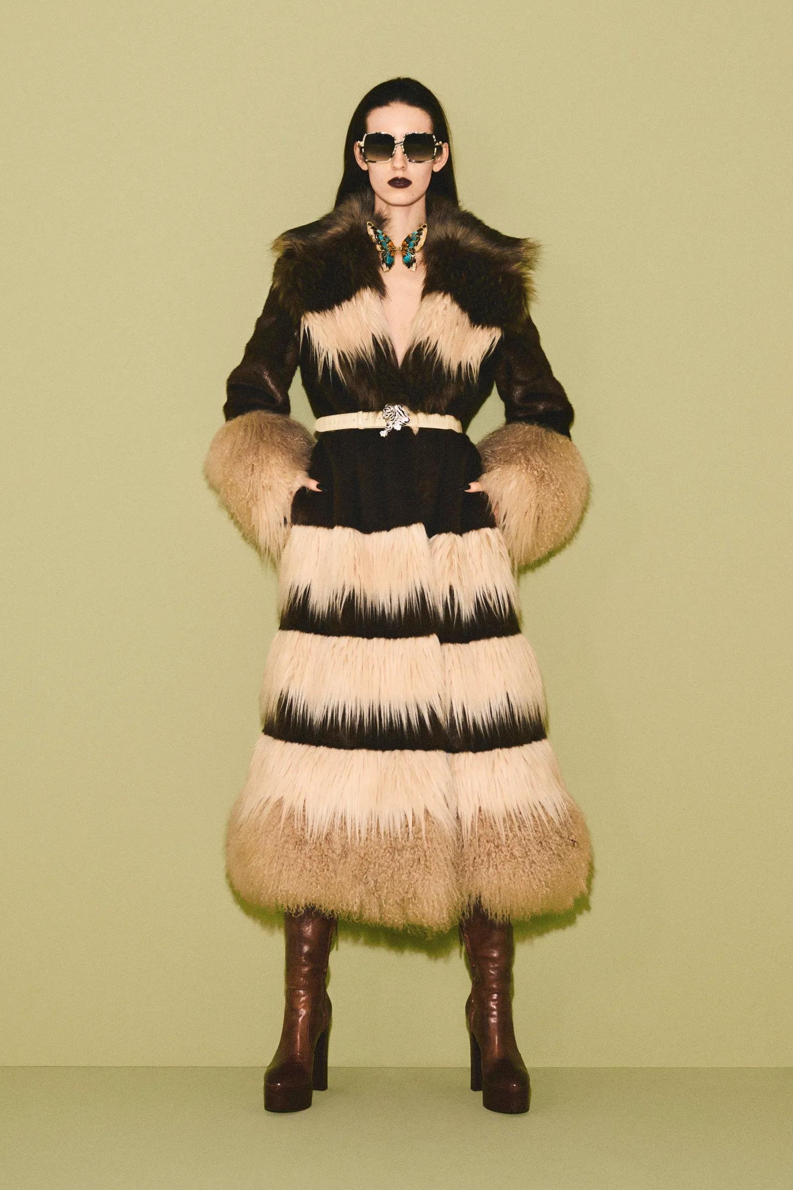 Woman wearing a long fur coat with beige, black, and dark brown stripes, dark sunglasses, dark lipstick, a butterfly necklace, and brown high-heeled boots.