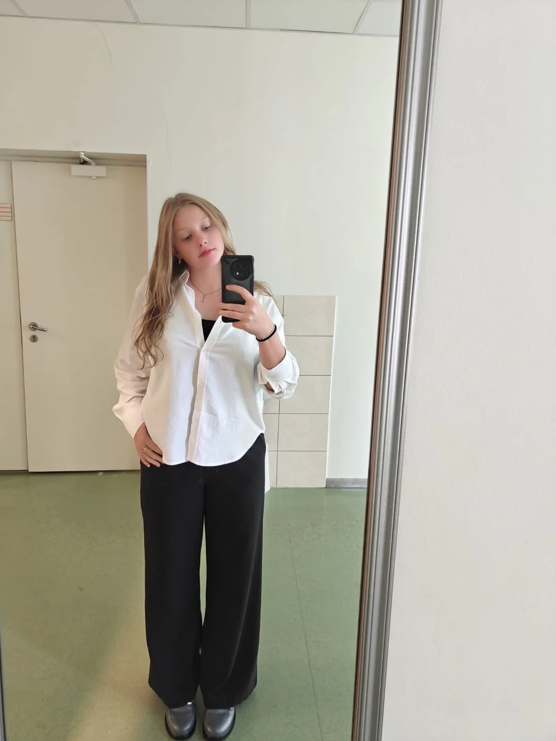 Woman taking a mirror selfie in a restroom, wearing a white button-up shirt, black pants, and metallic shoes.