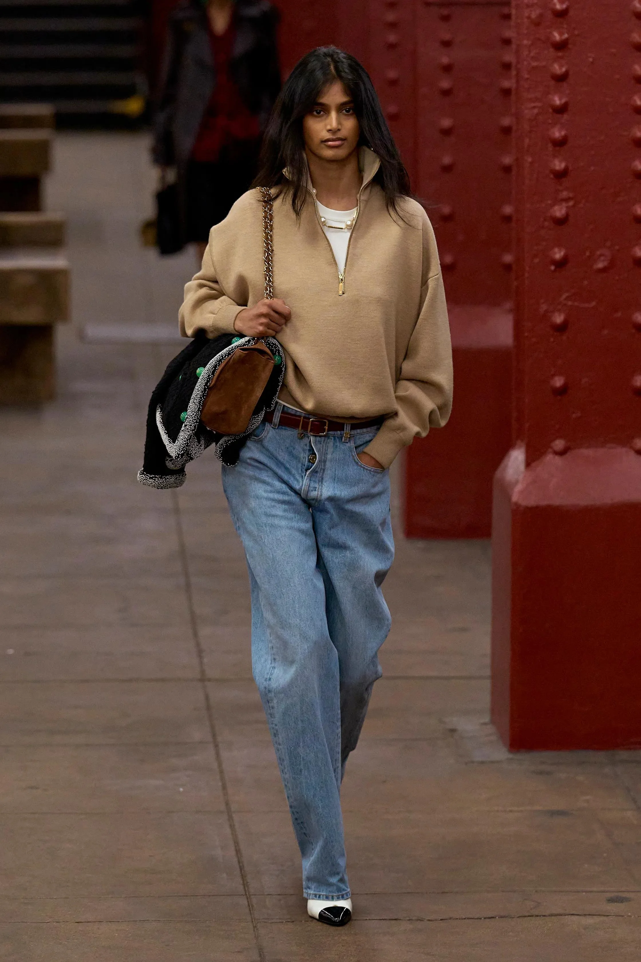 A woman with long black hair wearing a beige quarter-zip pullover, oversized blue jeans, and white shoes with black accents, walking indoors with her left hand in her pocket, carrying a black bag with a chain strap and a brown pouch.