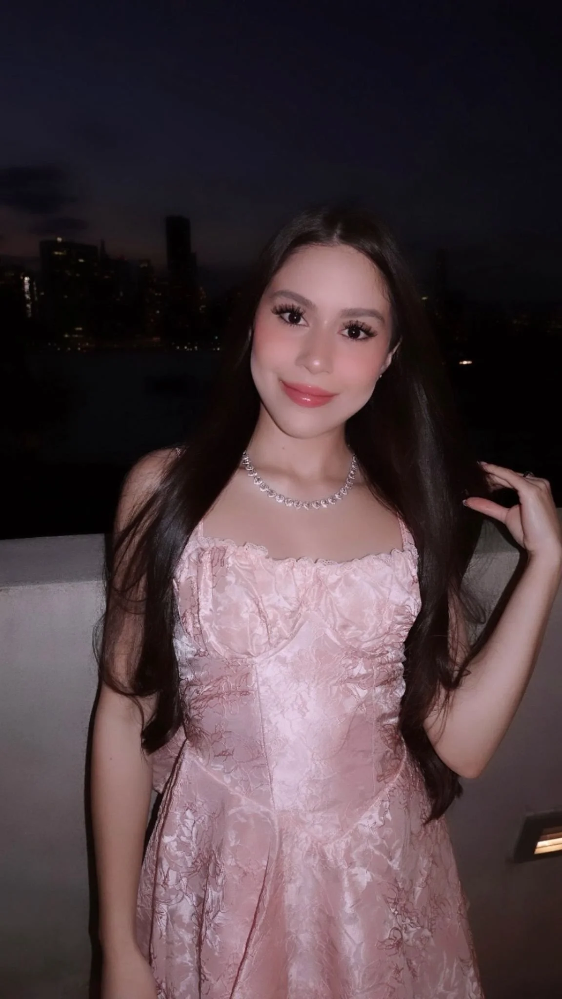 A young woman in a pink dress and a diamond necklace standing outdoors at night with a city skyline in the background.