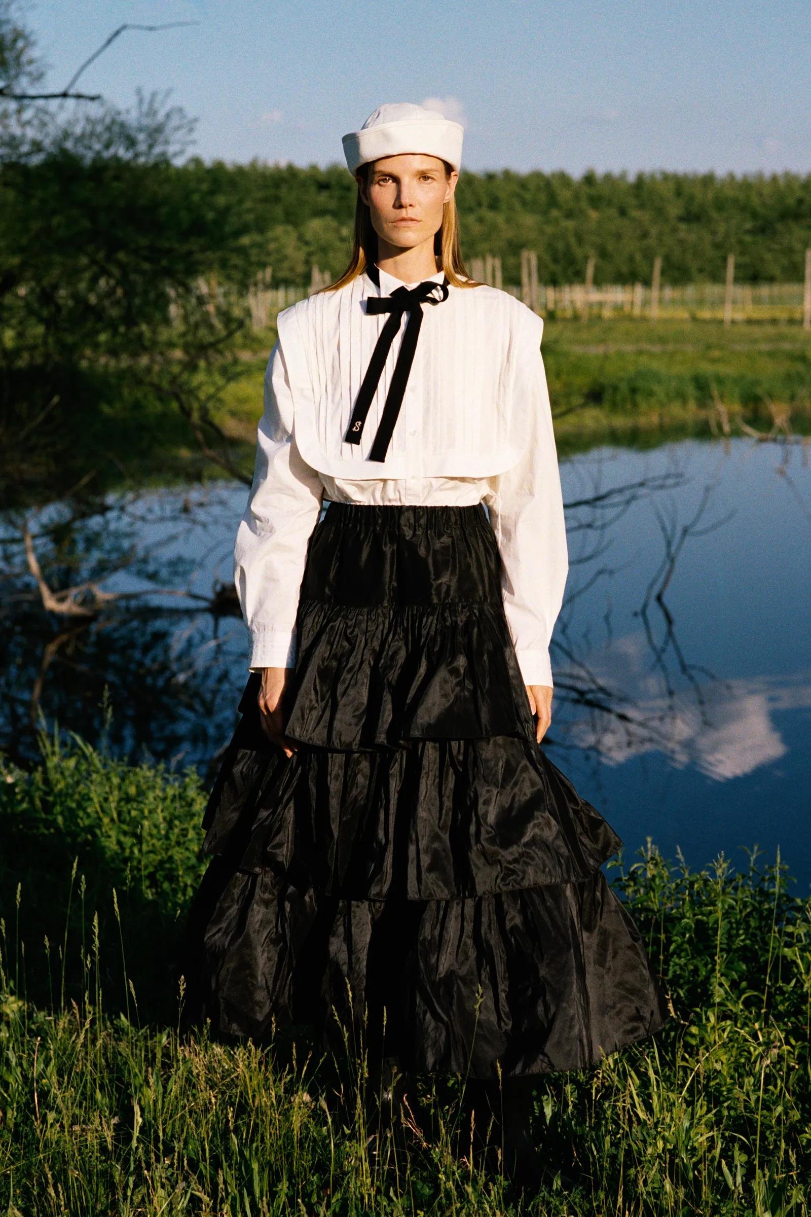 A woman standing outdoors by a body of water, wearing a white blouse with a black bow, black tiered skirt, and a white sailor-style hat, with green trees and a blue sky in the background.