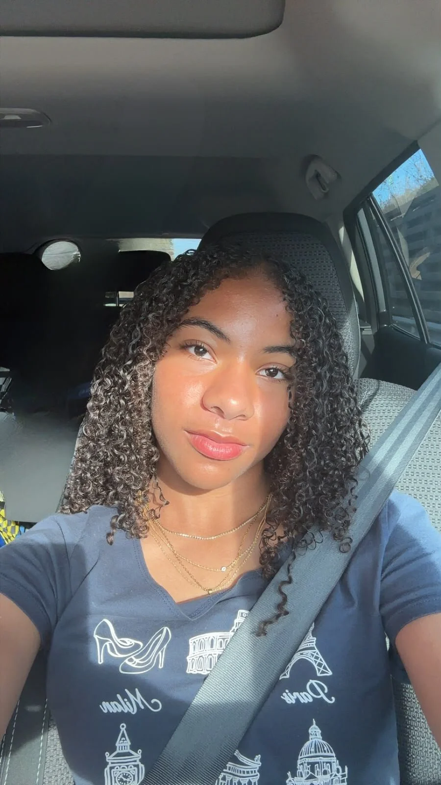 A young woman with curly hair taking a selfie inside a car, wearing a navy blue T-shirt with white illustrations of famous landmarks and gold jewelry, with sunlight illuminating her face.