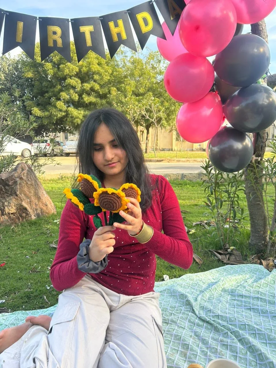 A young girl at an outdoor birthday party sitting on a picnic blanket, holding crochet sunflower and leaf decorations, with pink and black balloons and a black pennant banner with yellow letters spelling 'HAPPY BIRTHDAY' in the background, trees and parked cars are also visible.
