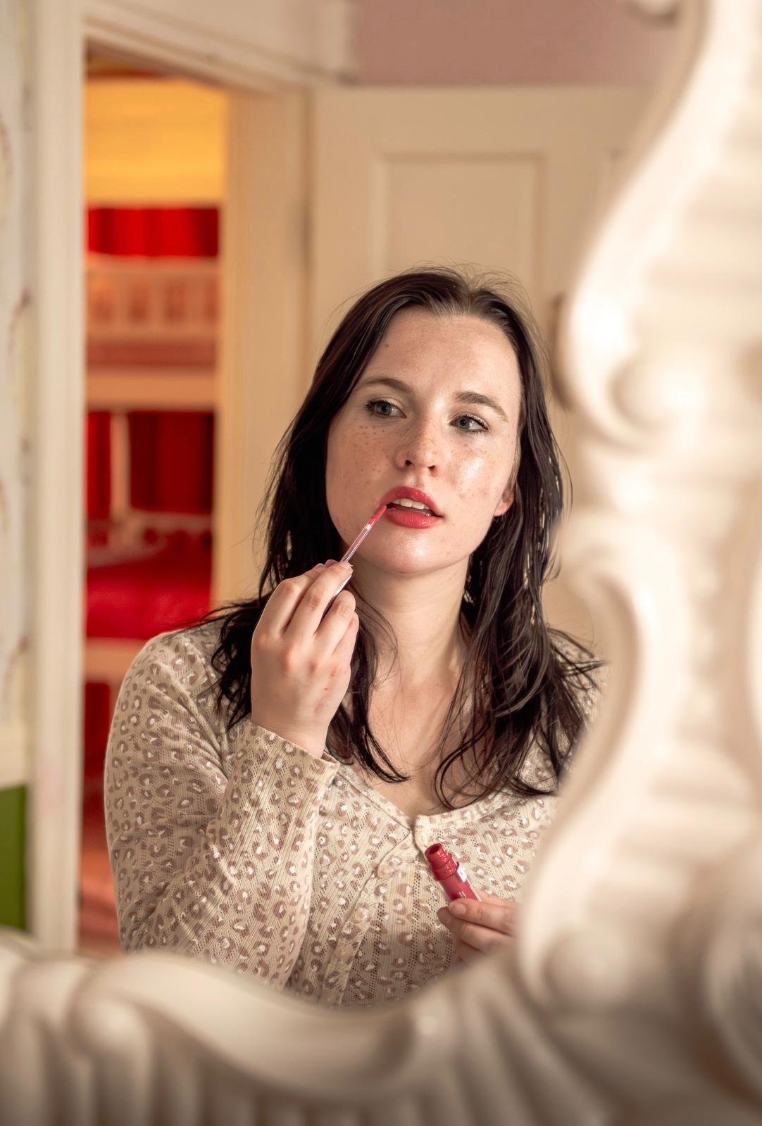 A woman with dark hair and freckles looking into a mirror, applying lipstick using a lip gloss applicator, in a room with warm lighting and decorative furniture.