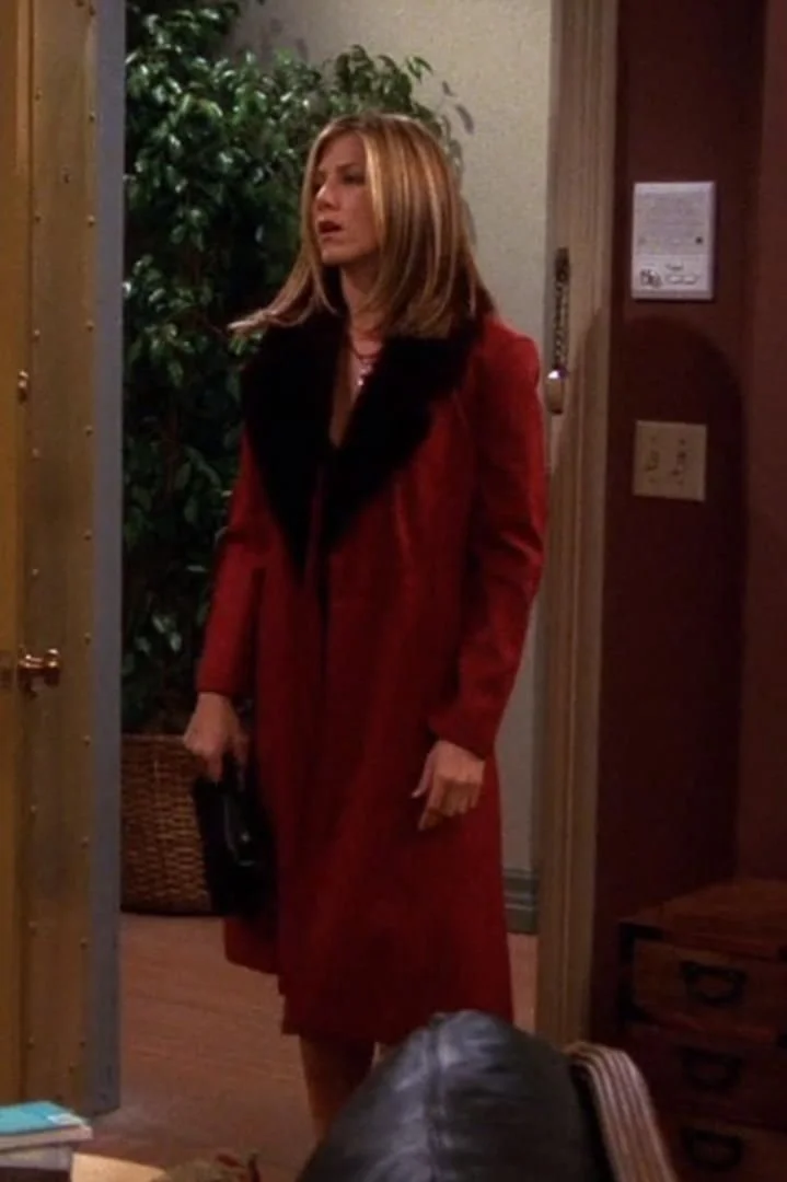 A woman with shoulder-length blonde hair wearing a red coat with a black fur collar, holding a small black purse, standing inside a house near the doorway.