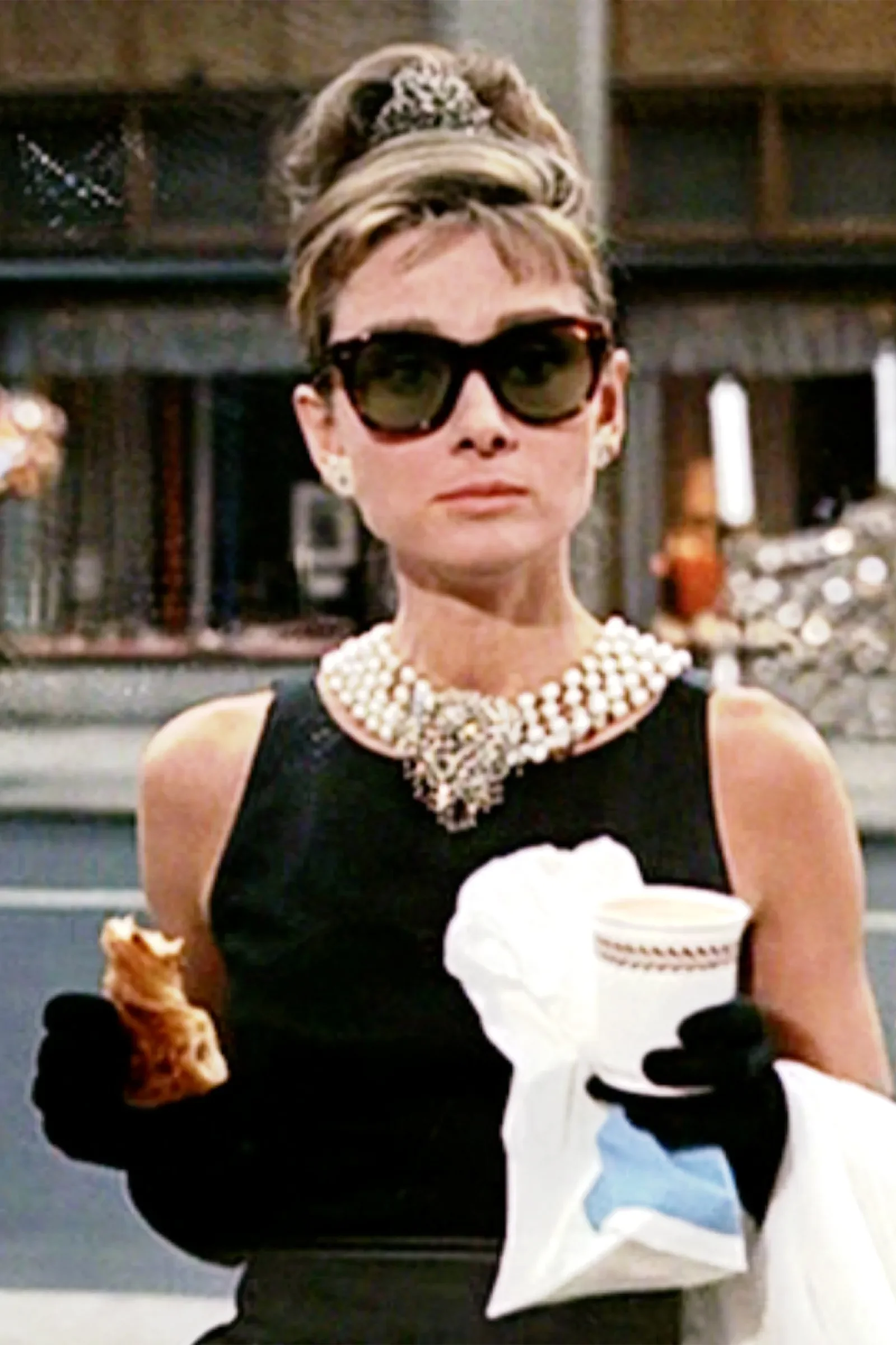 A woman with short hair, wearing large sunglasses, a multi-strand pearl necklace, black dress, and black gloves, holding a white napkin and a cup, in a possibly urban or store setting with shelves in the background.