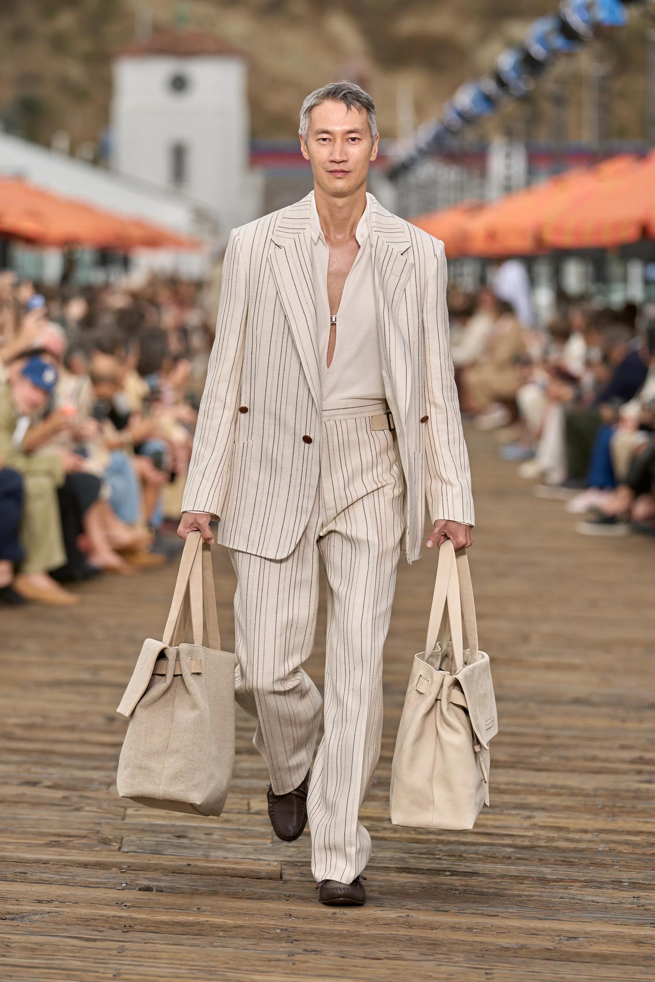 A male model walking on a wooden runway during a fashion show, carrying beige tote bags in each hand, dressed in a cream-colored pinstripe suit with a partially unbuttoned shirt.