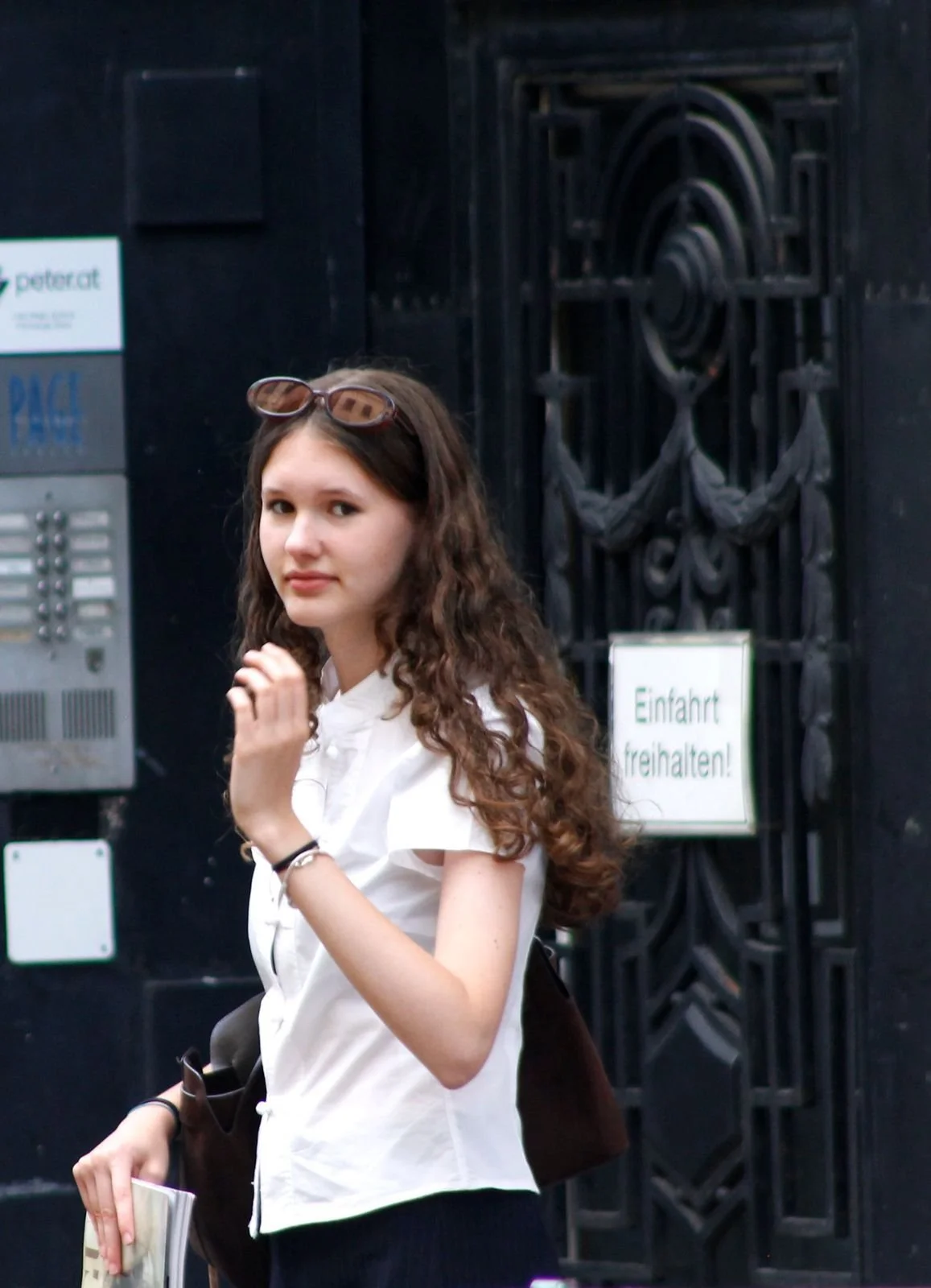 Young woman with curly brown hair, wearing sunglasses on her head and a white shirt, standing near a black gate with signs, holding a booklet or magazine.