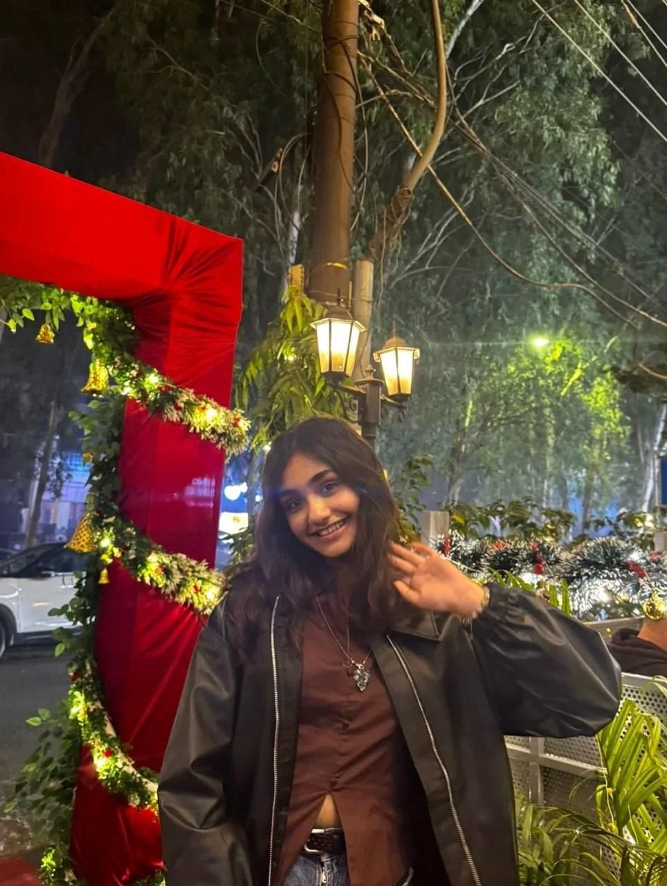 A young woman with dark hair smiling and waving at the camera while standing outdoors at night, surrounded by festive holiday decorations including a red and green wreath, string lights, and trees, with a street scene in the background.