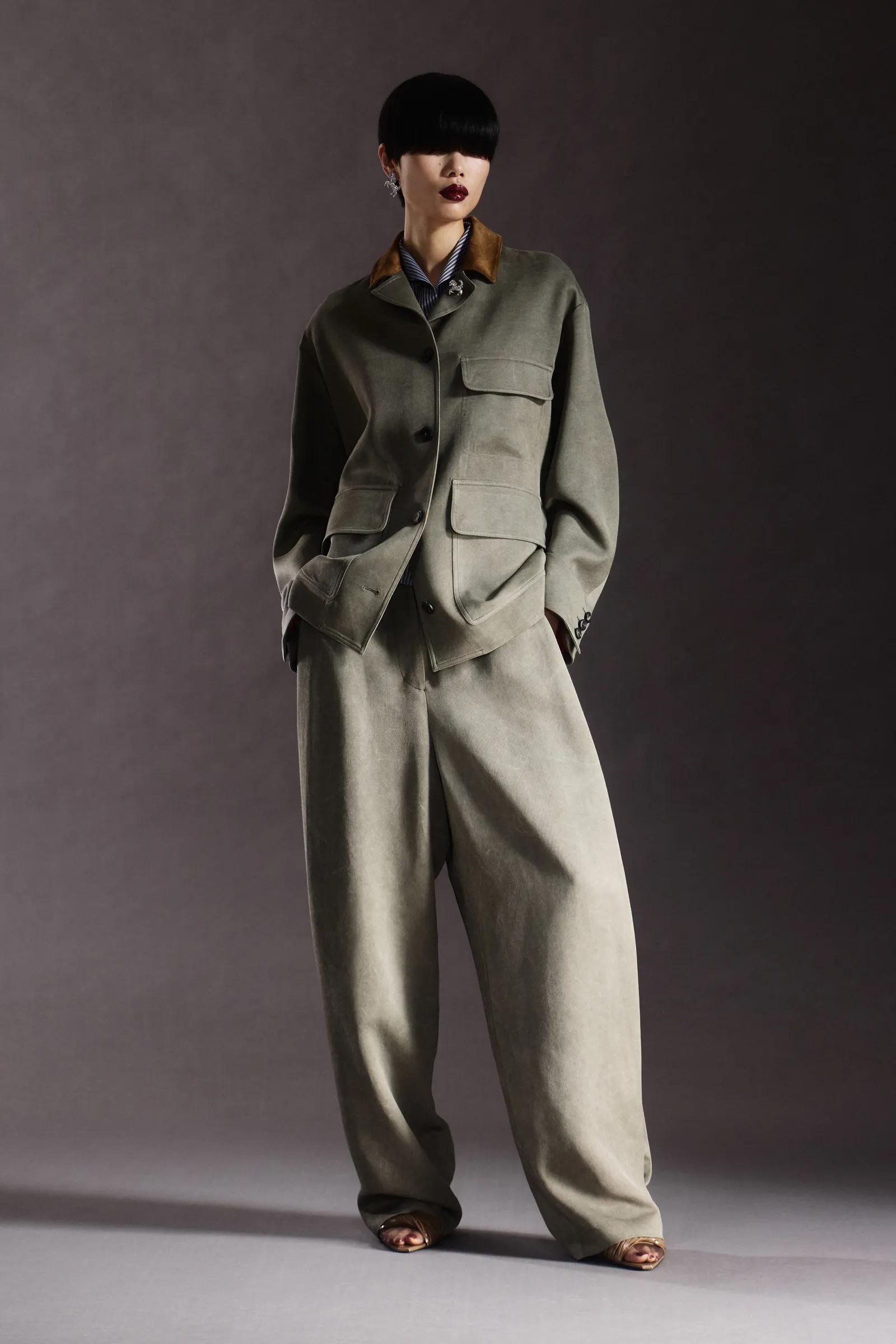 Fashion model wearing a beige oversized suit and dark lipstick, standing against a dark background.