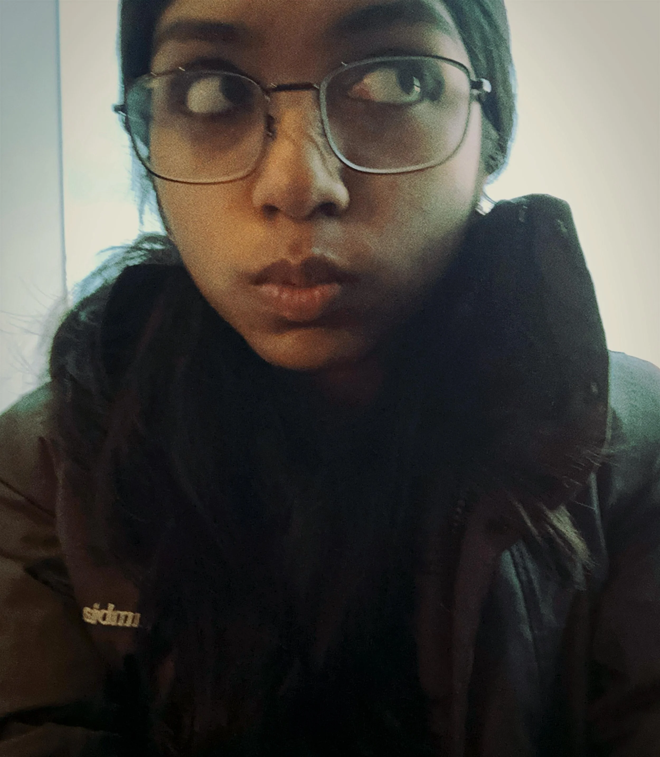 Close-up of a person's face with glasses, dark hair, and wearing a black hoodie with a visible logo.