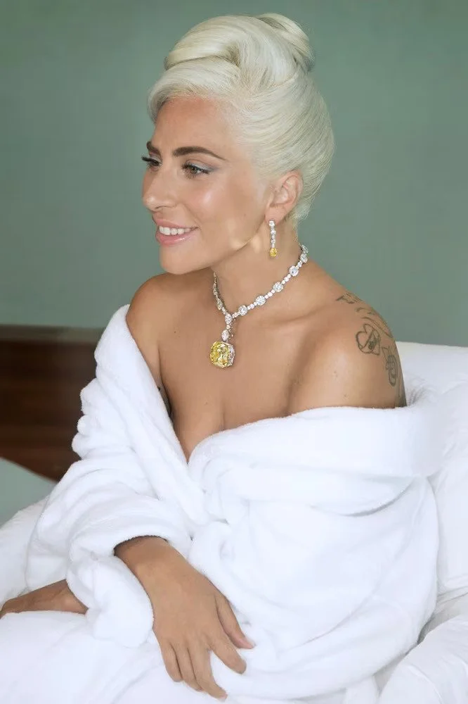 A woman with platinum blonde hair styled in an elegant updo, wearing diamond jewelry with a large yellow gemstone pendant, sitting on a bed with a white robe, smiling softly, against a muted green background.