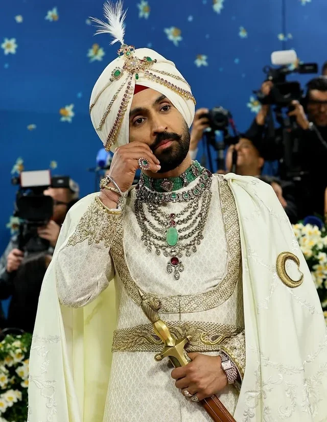 A man dressed as a prince or king wearing white and gold traditional Indian attire with a turban, jewelry, and a sword, surrounded by photographers in a setting with a blue background and yellow flowers.