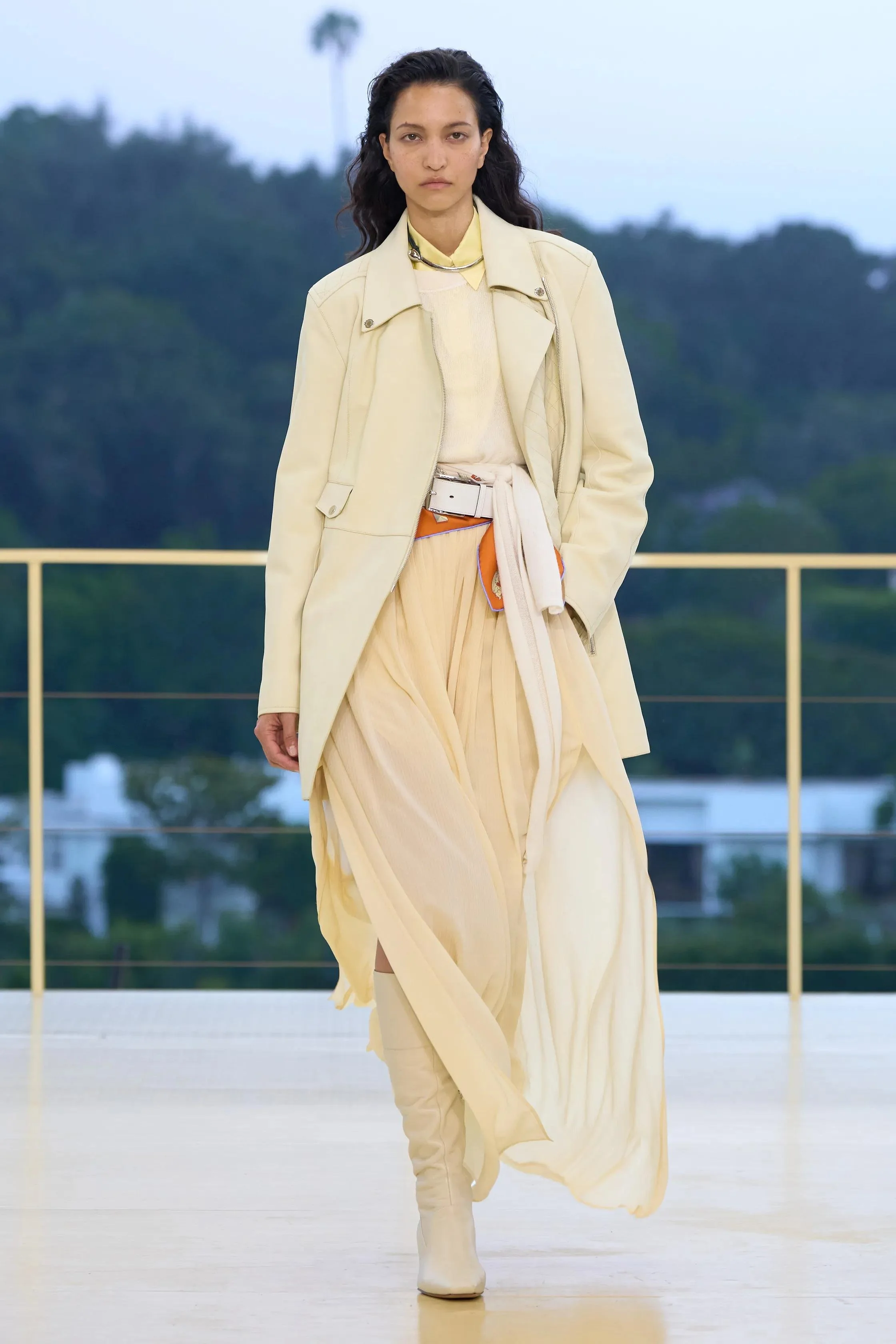 A woman walking outdoors on a balcony, dressed in beige and cream fashion, with a backdrop of trees and a cloudy sky.