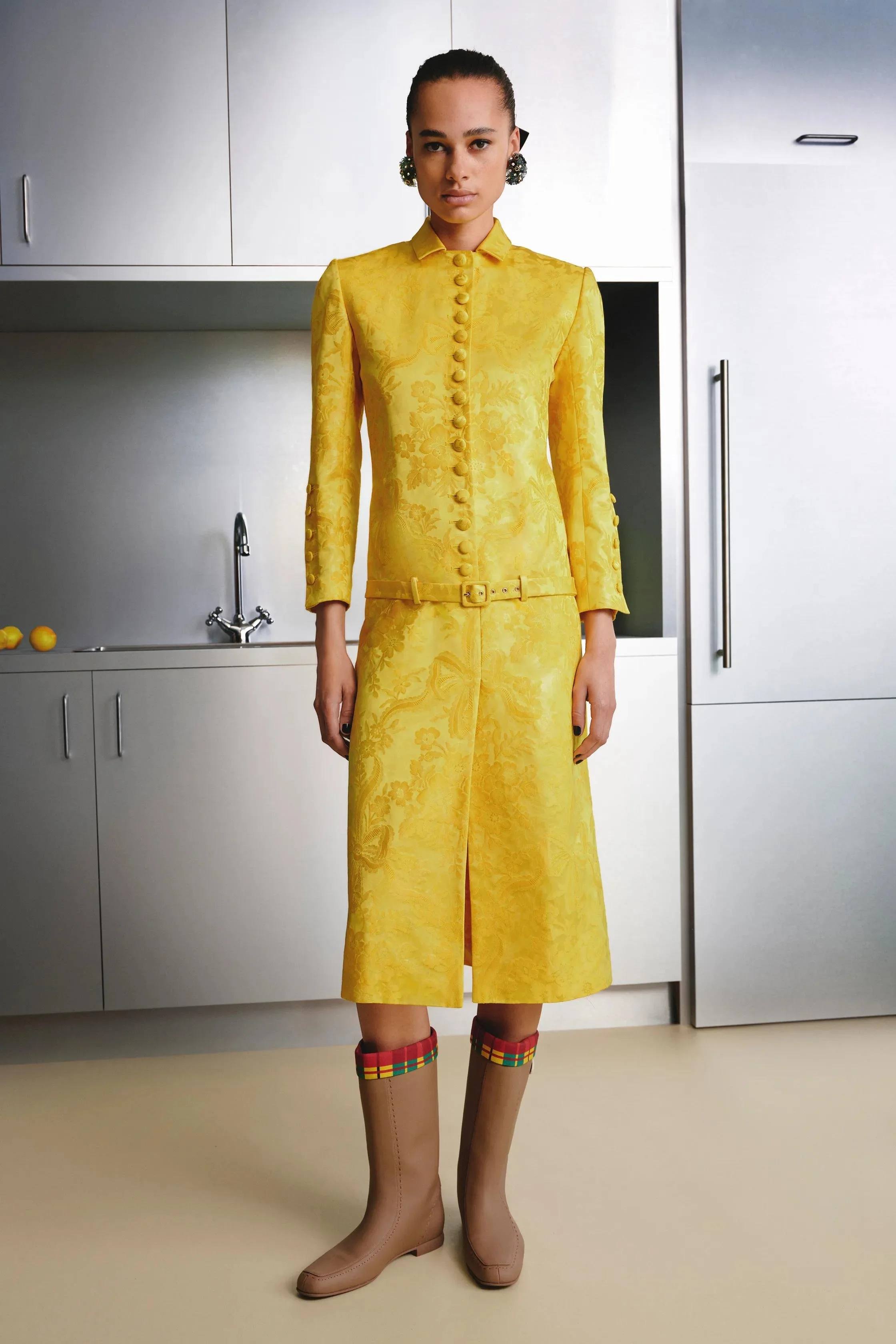 A woman standing in a kitchen wearing a yellow floral three-piece suit with matching wide-leg pants, pink knee-high boots with multicolored plaid trim, and large floral earrings.
